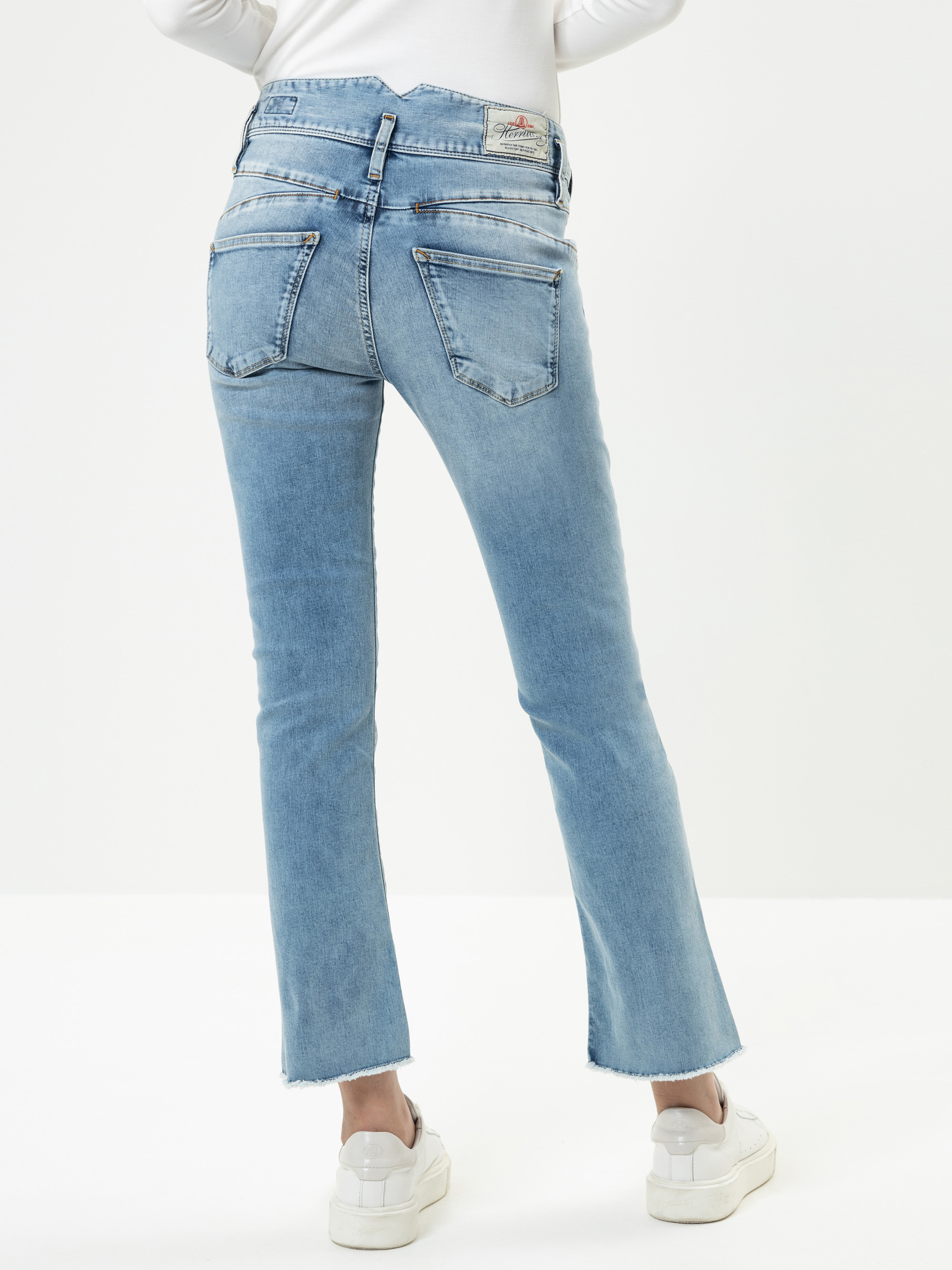 Eine Person, die eine hellblaue Jeans mit geradem Bein und ausgefransten Säumen und weiße Turnschuhe trägt, wird von hinten gezeigt, vor einem schlichten weißen Hintergrund stehend.