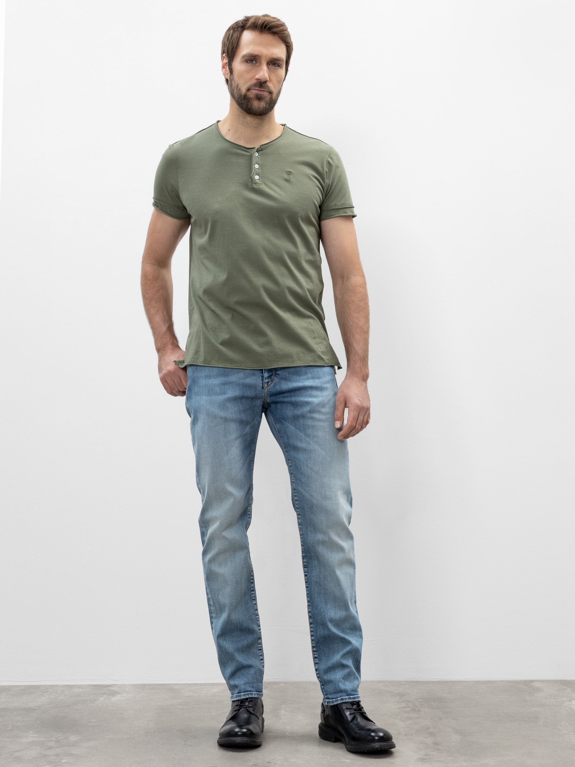 Ein Mann mit kurzen braunen Haaren und Bart steht vor einer schlichten weißen Wand. Er trägt ein grünes Henley-T-Shirt, hellblaue Jeans und schwarze Schuhe. Er hat einen neutralen Gesichtsausdruck und seine Arme liegen entspannt an seinen Seiten.