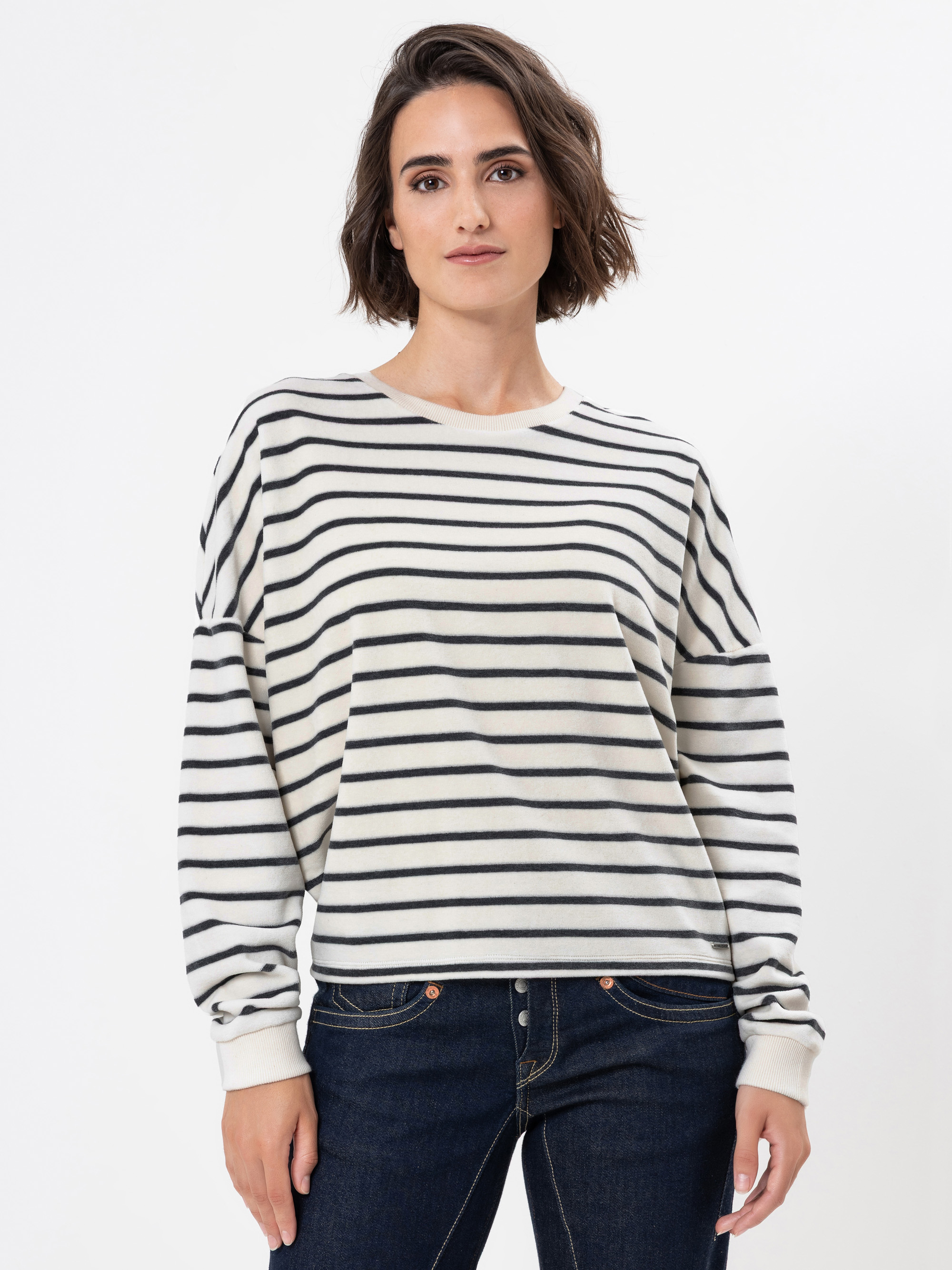Eine Frau mit kurzen braunen Haaren trägt einen weißen Pullover mit schwarzen Querstreifen und blaue Jeans und steht vor einem schlichten weißen Hintergrund.