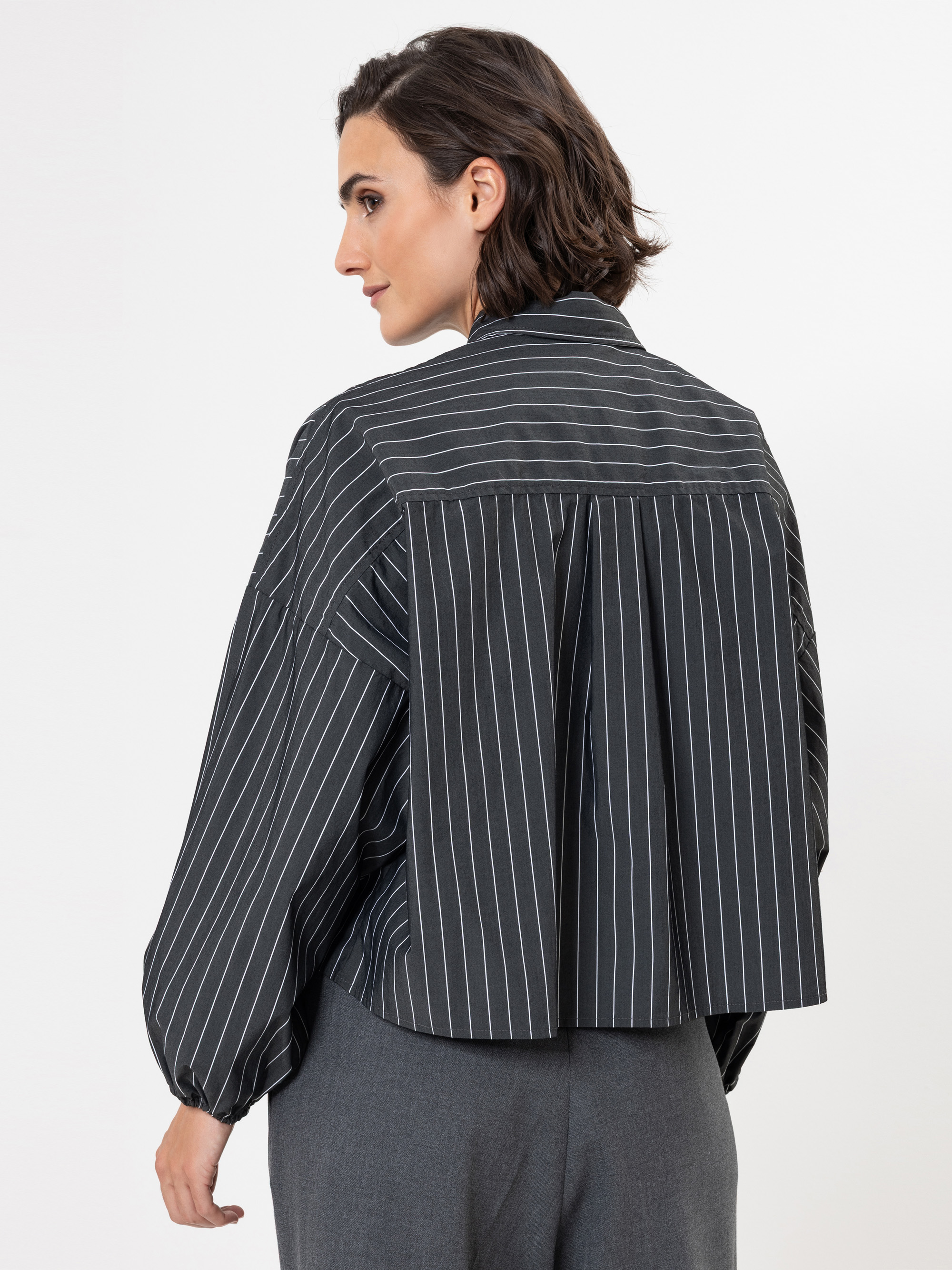 Eine Frau mit kurzen dunklen Haaren trägt eine locker sitzende, dunkle Nadelstreifenbluse mit langen Ärmeln und eine graue Hose. Sie steht vor einem schlichten weißen Hintergrund und blickt nach links.
