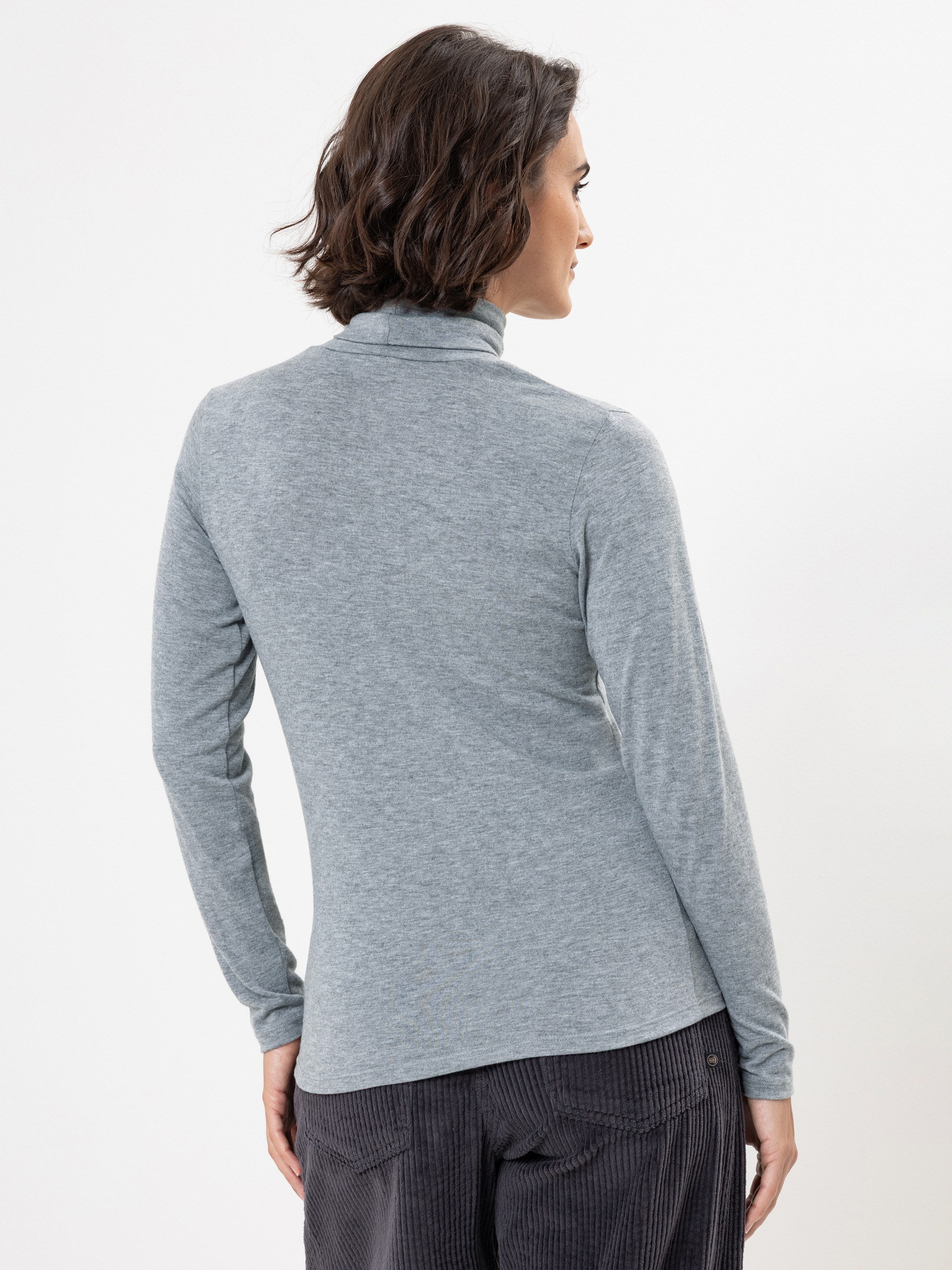 Eine Person mit kurzem, dunklem, gewelltem Haar blickt von der Kamera weg und trägt ein hellgraues, langärmeliges Rollkragenoberteil und eine dunkle Cordhose vor einem schlichten weißen Hintergrund.