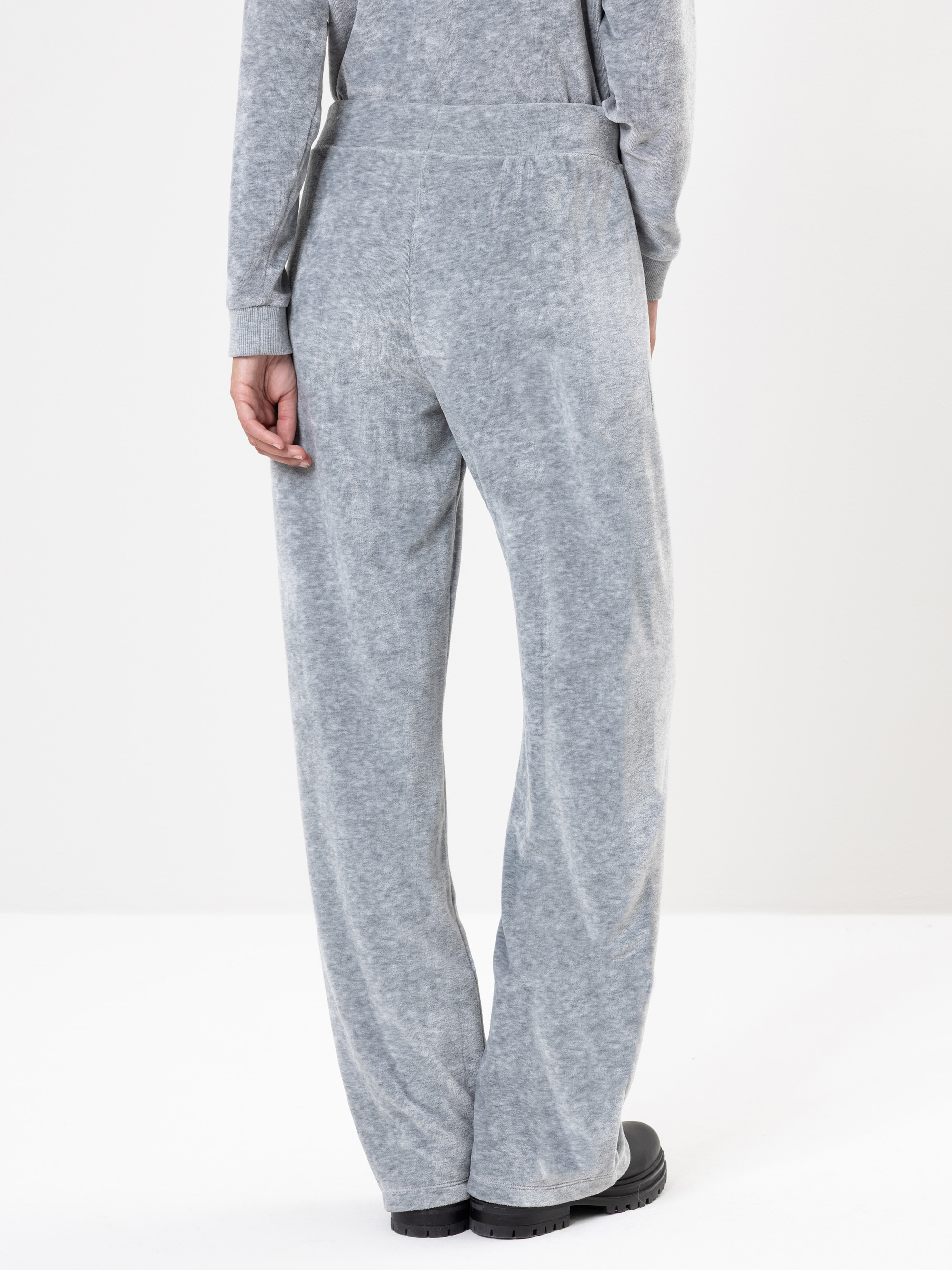 Stehende Person vor weißem Hintergrund, bekleidet mit einem hellgrauen, locker sitzenden, langärmeligen Oberteil und einer passenden Hose mit weitem Bein, dazu schwarze Schuhe. Das Bild ist von hinten aufgenommen und zeigt entspannten Loungewear-Stil.