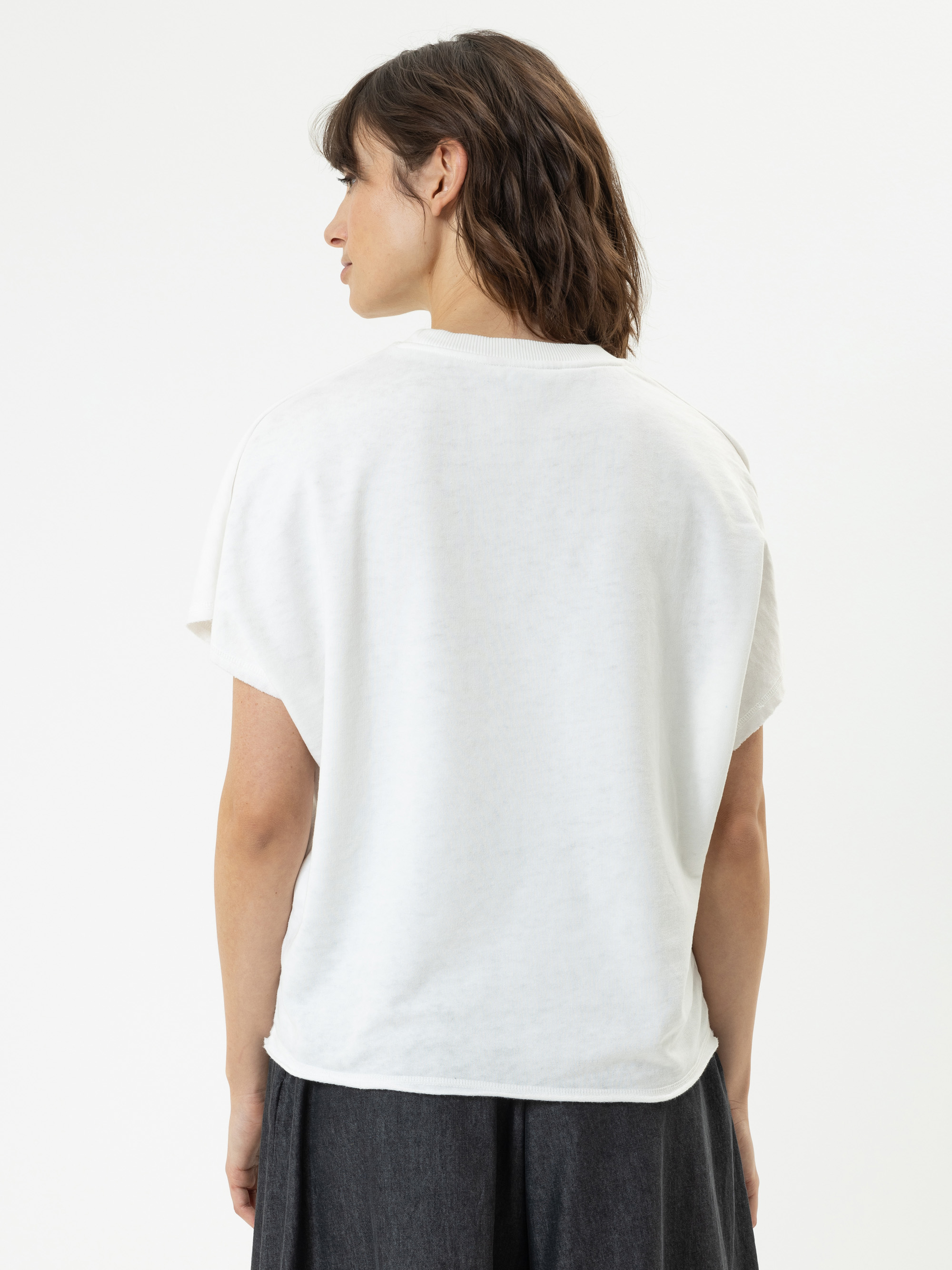 Eine Person mit kurzen braunen Haaren, von hinten gesehen, trägt ein lockeres weißes kurzärmeliges T-Shirt und eine dunkle Hose vor einem einfarbig weißen Hintergrund.