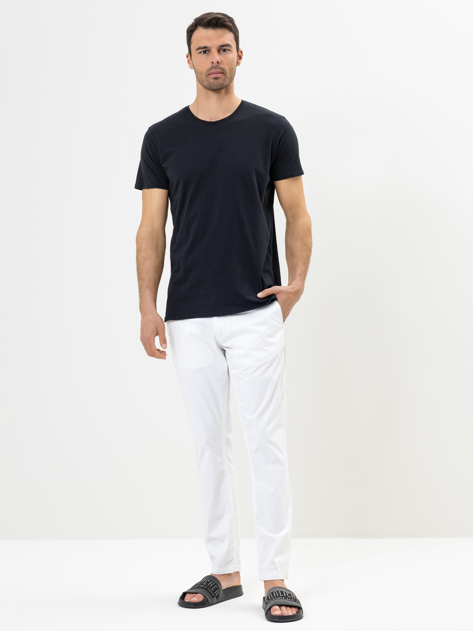 Ein Mann steht vor einem schlichten weißen Hintergrund und trägt ein schwarzes T-Shirt, eine weiße Hose und schwarze Slide-Sandalen. Eine Hand steckt in seiner Hosentasche und er blickt mit einem neutralen Gesichtsausdruck nach vorne.