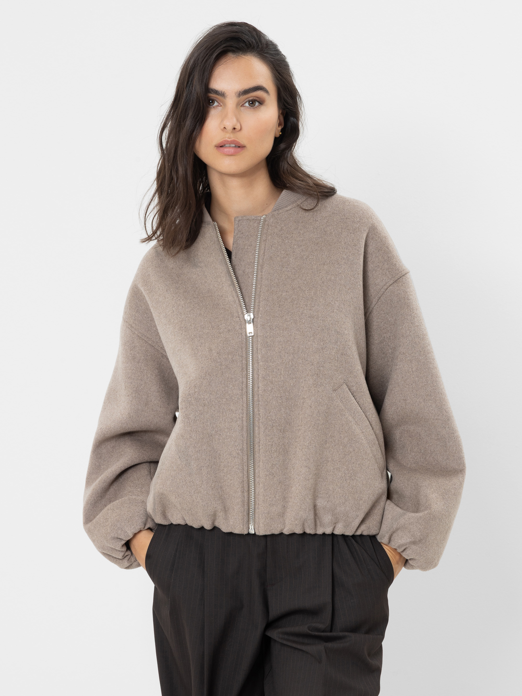 Eine Frau mit welligem, dunklem Haar trägt eine beige Jacke mit Reißverschluss, Seitentaschen und weiten Ärmeln sowie eine schwarze Hose und steht vor einem schlichten weißen Hintergrund.