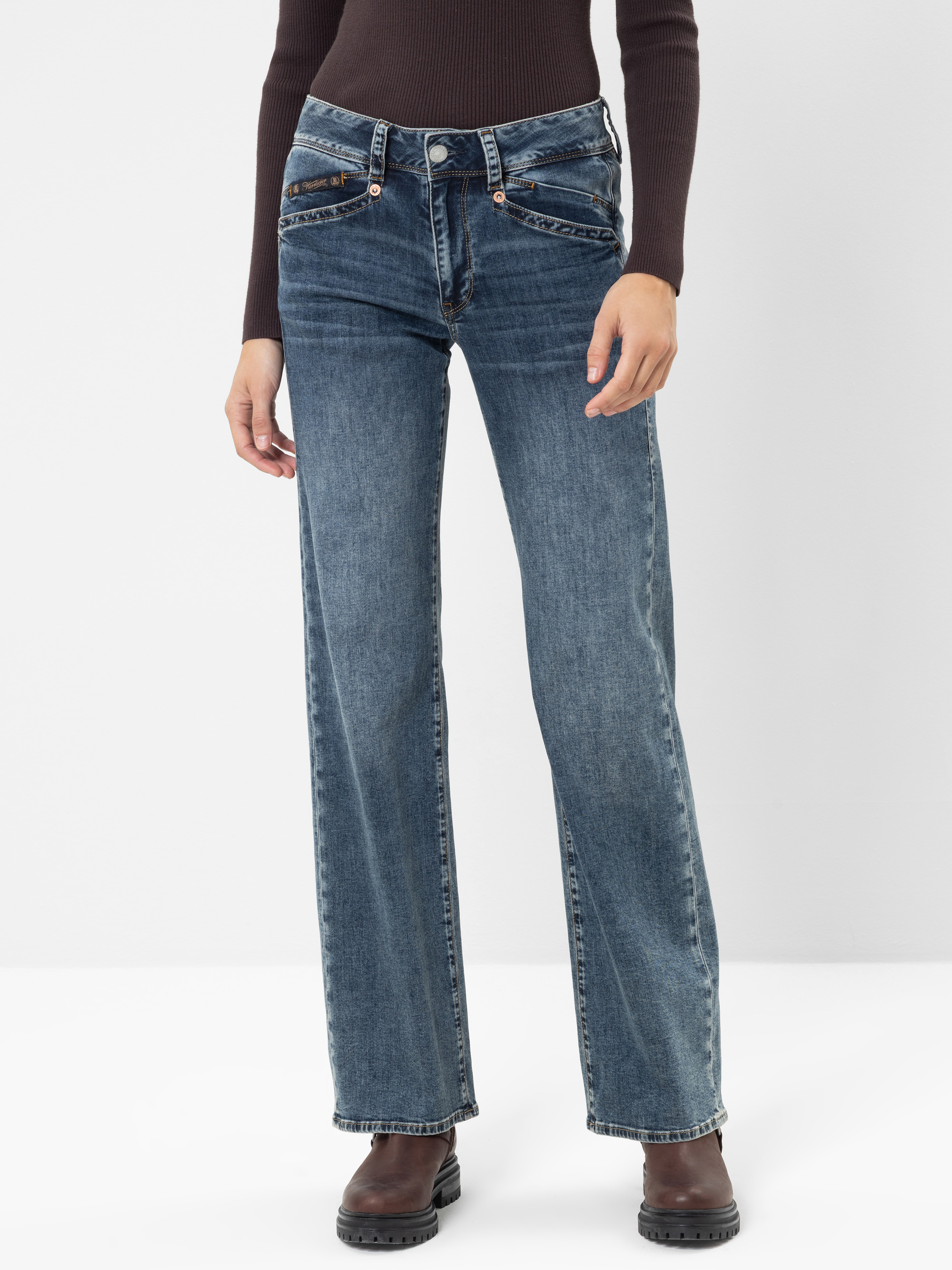 Eine Person, die eine blaue Jeans mit hoher Taille und geradem Bein, ein eng anliegendes, dunkles Langarm-Oberteil und braune Stiefeletten trägt, steht vor einem schlichten weißen Hintergrund.