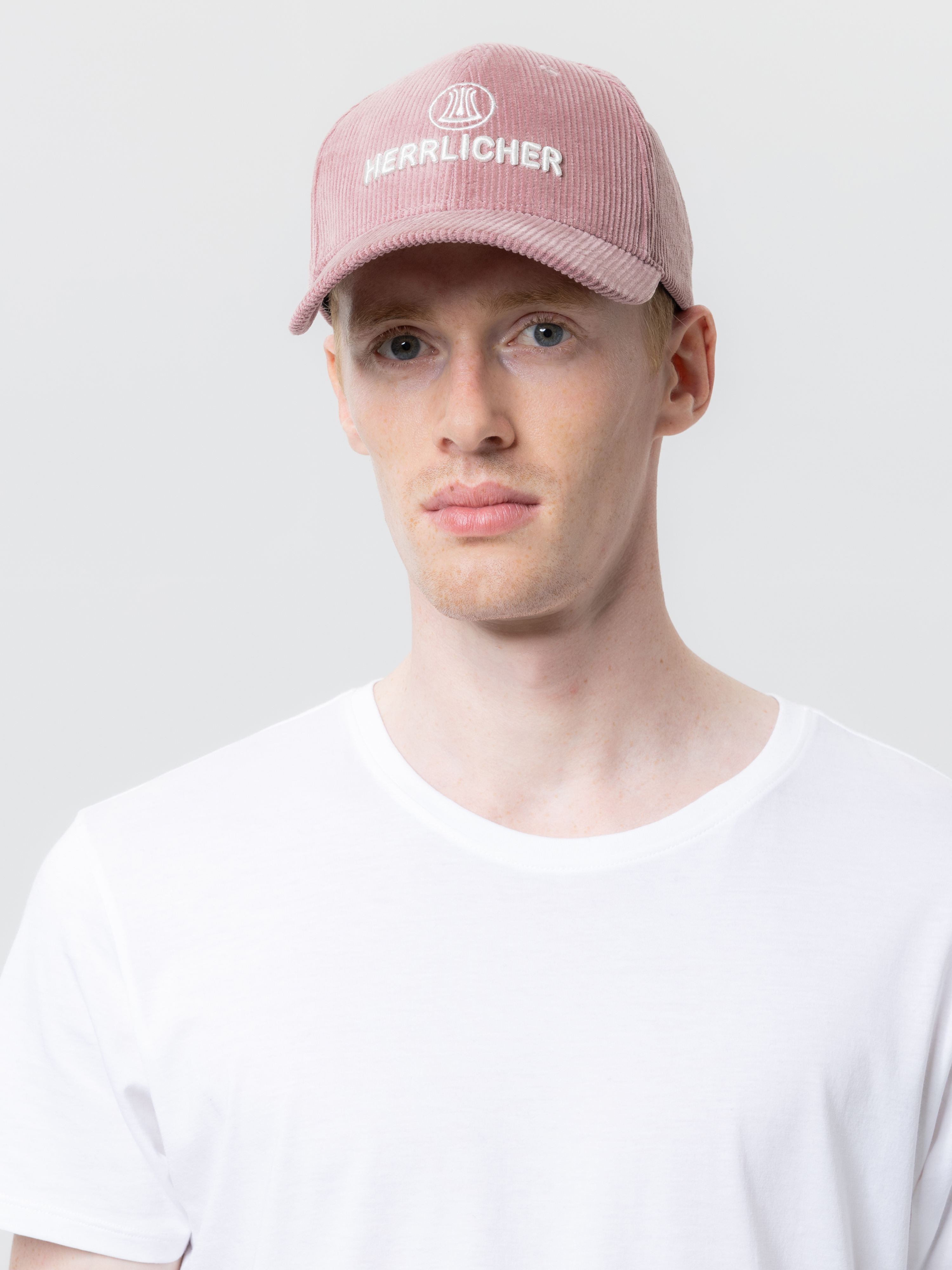 Eine Person, die eine blassrosa Kordmütze mit dem aufgestickten Wort HERRLICHER auf der Vorderseite und ein schlichtes weißes T-Shirt trägt, steht vor einem schlichten hellen Hintergrund.
