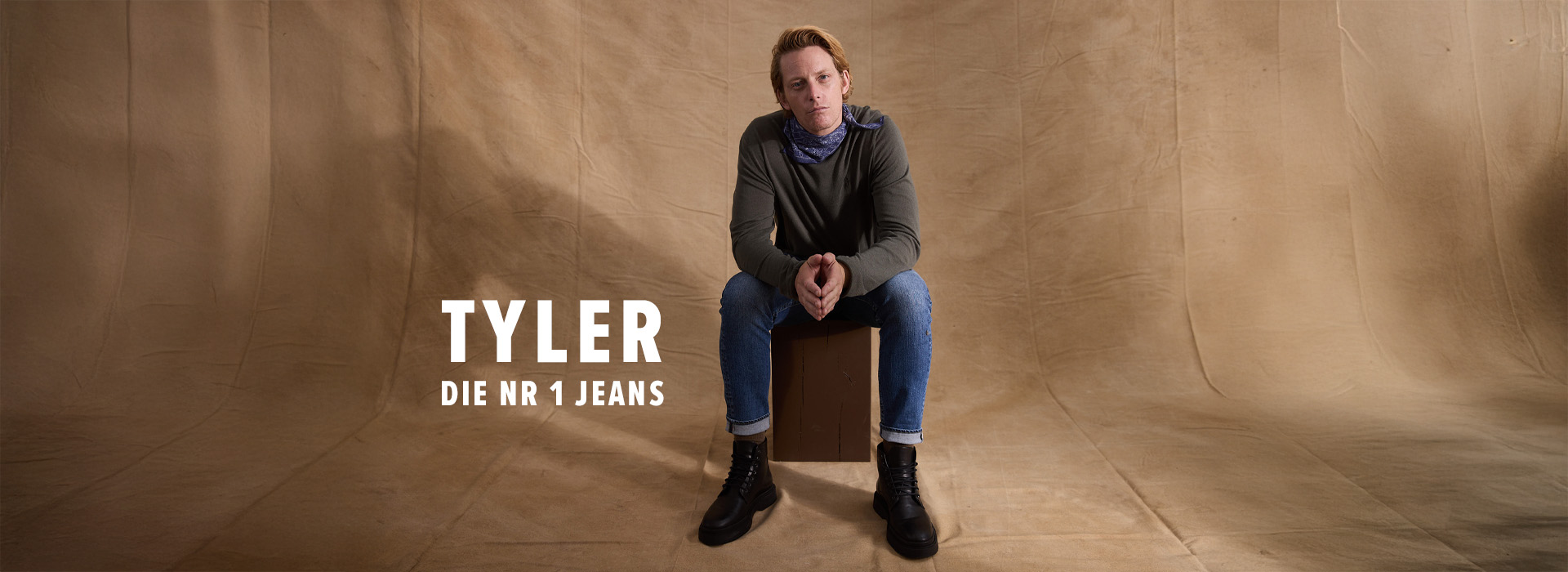 Ein Mann in einem grauen Hemd, blauen Jeans und schwarzen Stiefeln sitzt auf einer Kiste vor einem Hintergrund aus hellbraunem Stoff. Der weiße Text lautet TYLER DIE NR 1 JEANS.