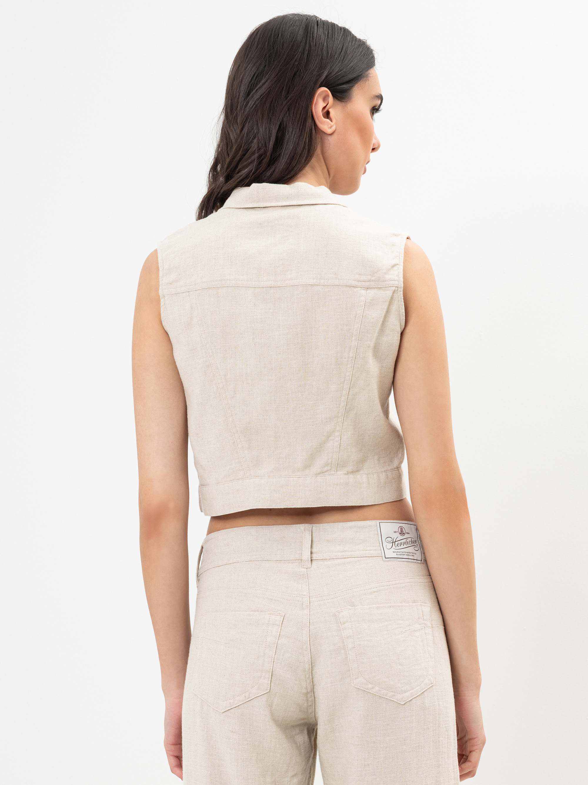 Eine Frau steht mit dem Gesicht zur Wand, trägt eine ärmellose beigefarbene Weste und eine dazu passende hochtaillierte Hose mit einem Aufnäher am Bund vor einem schlichten weißen Hintergrund.