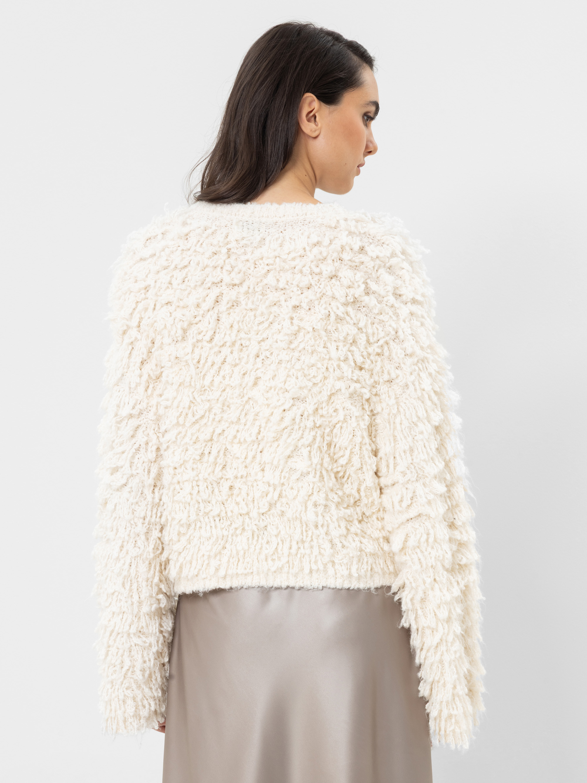 Eine Frau mit langen dunklen Haaren trägt einen strukturierten, cremefarbenen, flauschigen Pullover und einen seidigen, hellgrauen Rock. Sie steht mit dem Rücken zur Kamera und präsentiert die Rückseite ihres Outfits vor einem schlichten weißen Hinte