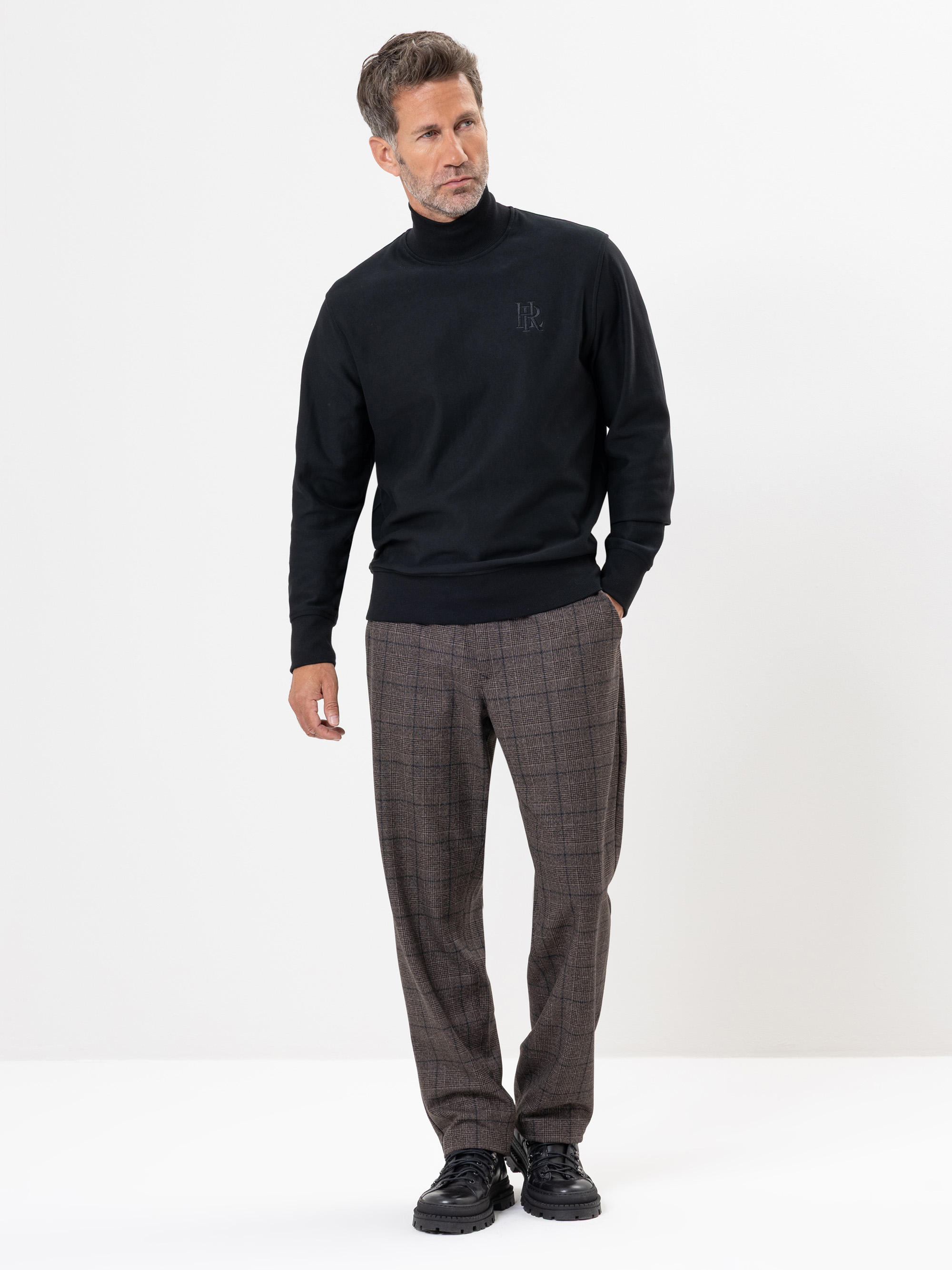Ein Mann steht vor einem weißen Hintergrund, trägt einen schwarzen Rollkragenpullover, eine braun karierte Hose und schwarze Schuhe, hat eine Hand in der Tasche und schaut leicht zur Seite.