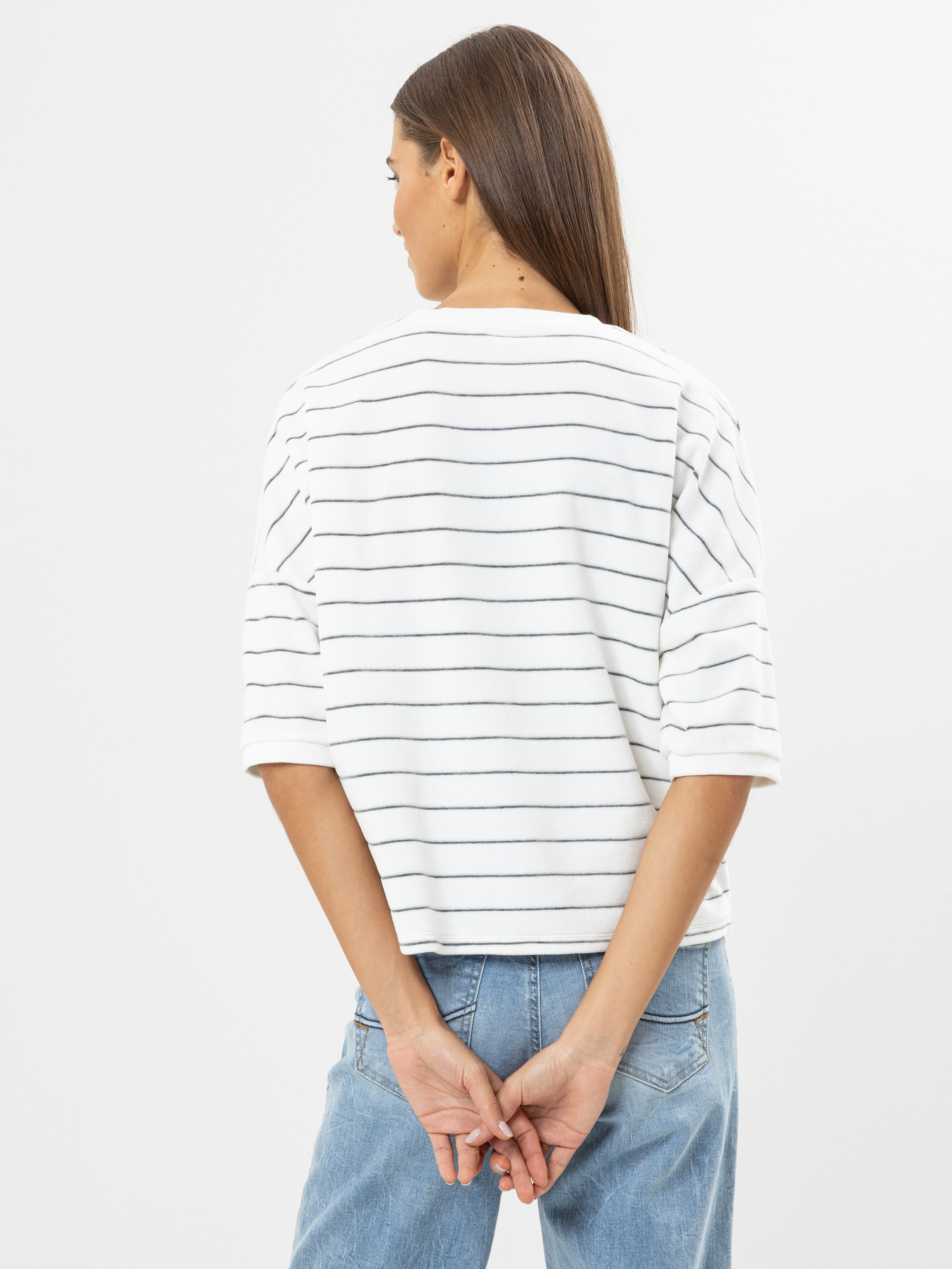 Eine Frau mit langen braunen Haaren steht mit dem Gesicht zur Wand, trägt ein weißes Oberteil mit dünnen schwarzen Querstreifen und hellblaue Jeans. Ihre Hände sind locker hinter ihrem Rücken verschränkt. Der Hintergrund ist einfarbig weiß.