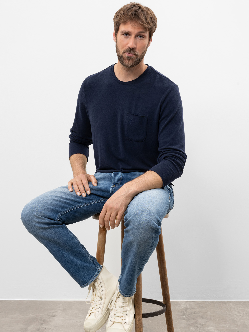 Ein Mann mit kurzen braunen Haaren und einem Bart sitzt auf einem Holzschemel vor einem schlichten weißen Hintergrund. Er trägt ein marineblaues langärmeliges Hemd, hellblaue Jeans und weiße Turnschuhe und blickt ruhig in die Kamera.