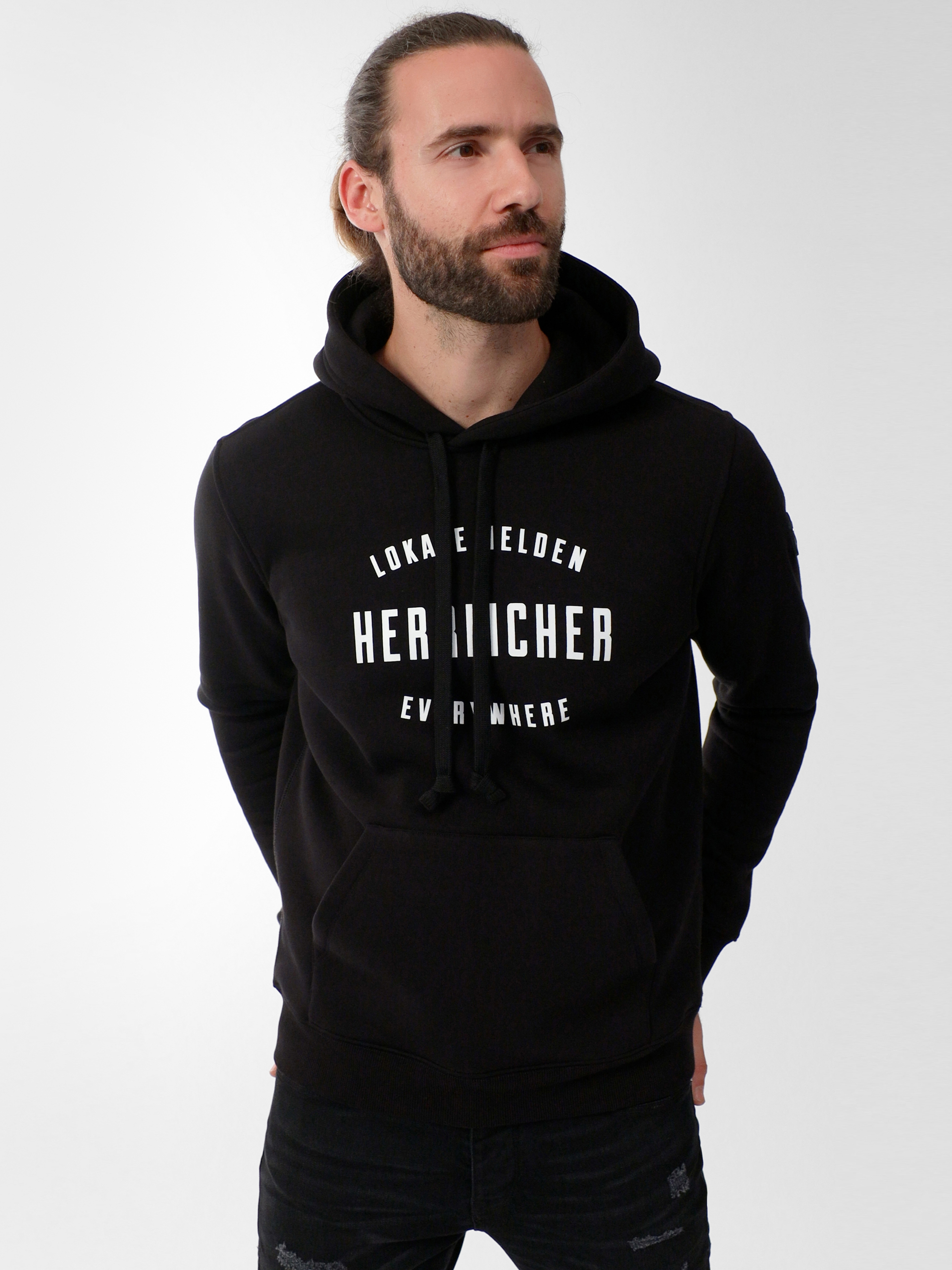 Ein Mann mit langen, zurückgebundenen Haaren und Bart trägt einen schwarzen Kapuzenpulli mit der weißen Aufschrift LOKA HELDEN HERHICHER ÜBERALL. Er steht mit den Händen in der Kapuzentasche vor einem schlichten hellgrauen Hintergrund.