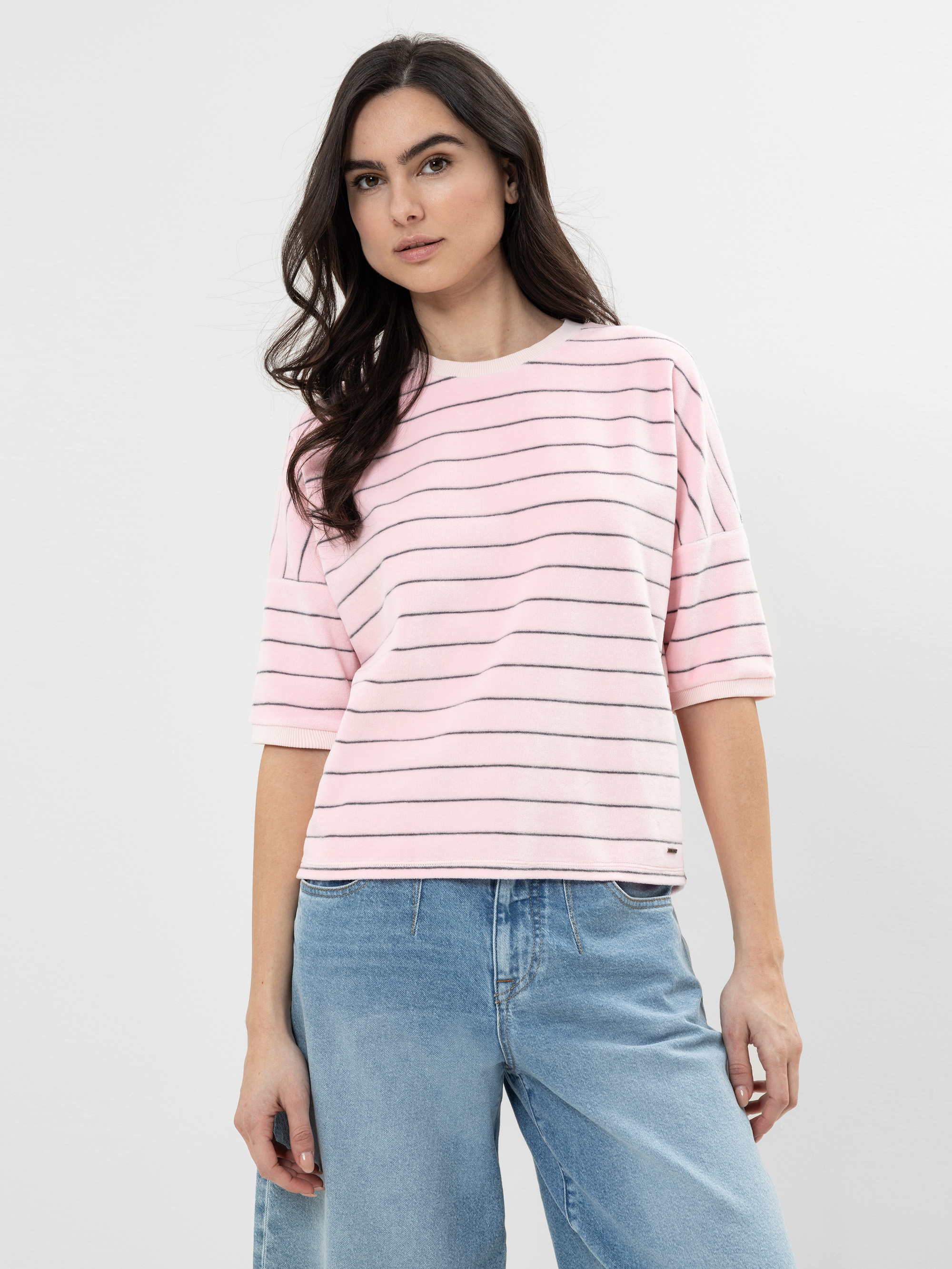 Eine Person mit langen dunklen Haaren trägt ein hellrosa, kurzärmeliges, gestreiftes Oberteil und blaue Jeans und steht vor einem schlichten hellen Hintergrund.
