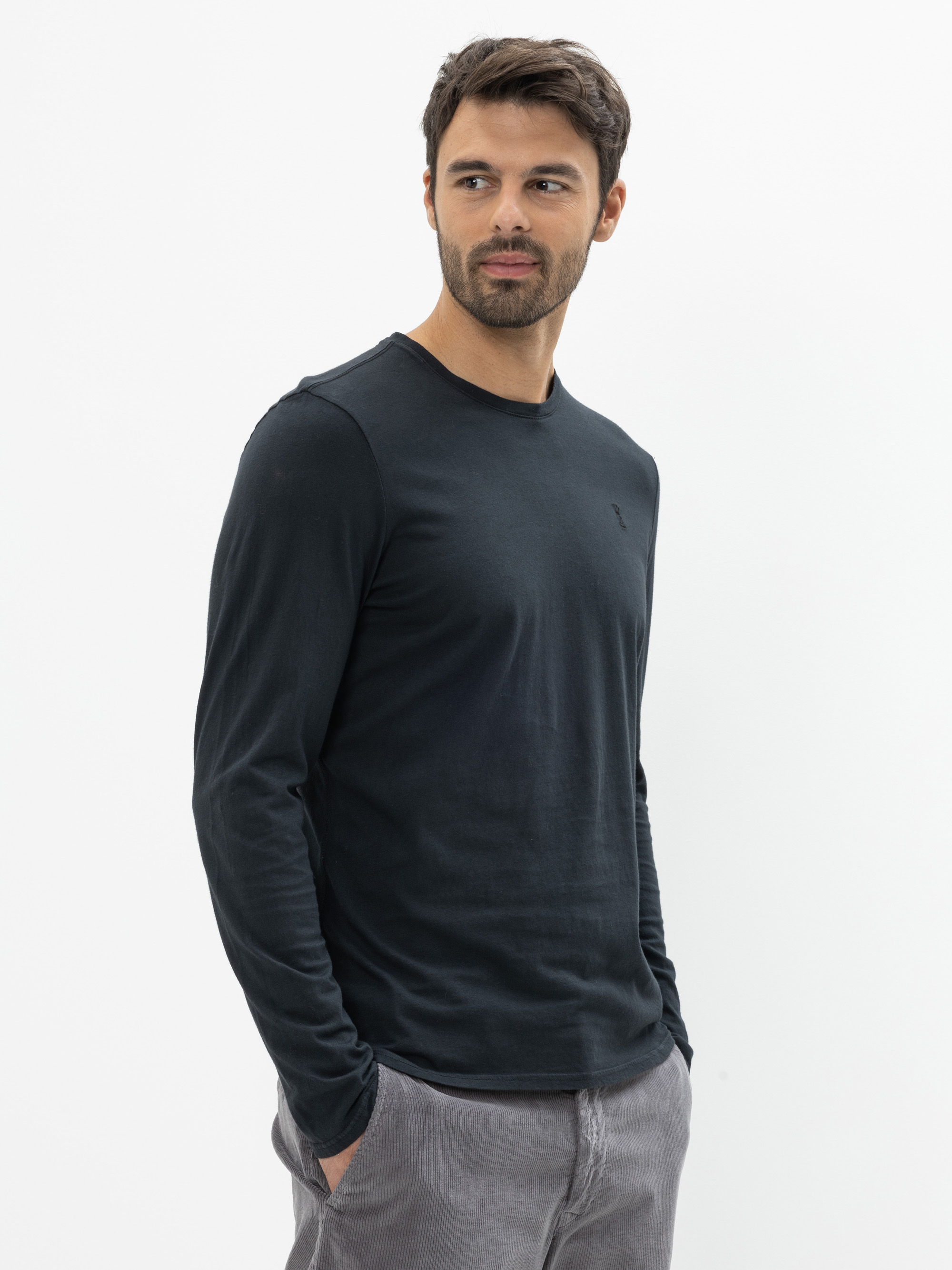 Ein Mann mit kurzen dunklen Haaren und einem Bart, der ein langärmeliges schwarzes Hemd und eine hellgraue Hose trägt, steht vor einem schlichten weißen Hintergrund und blickt mit neutraler Miene leicht nach links.