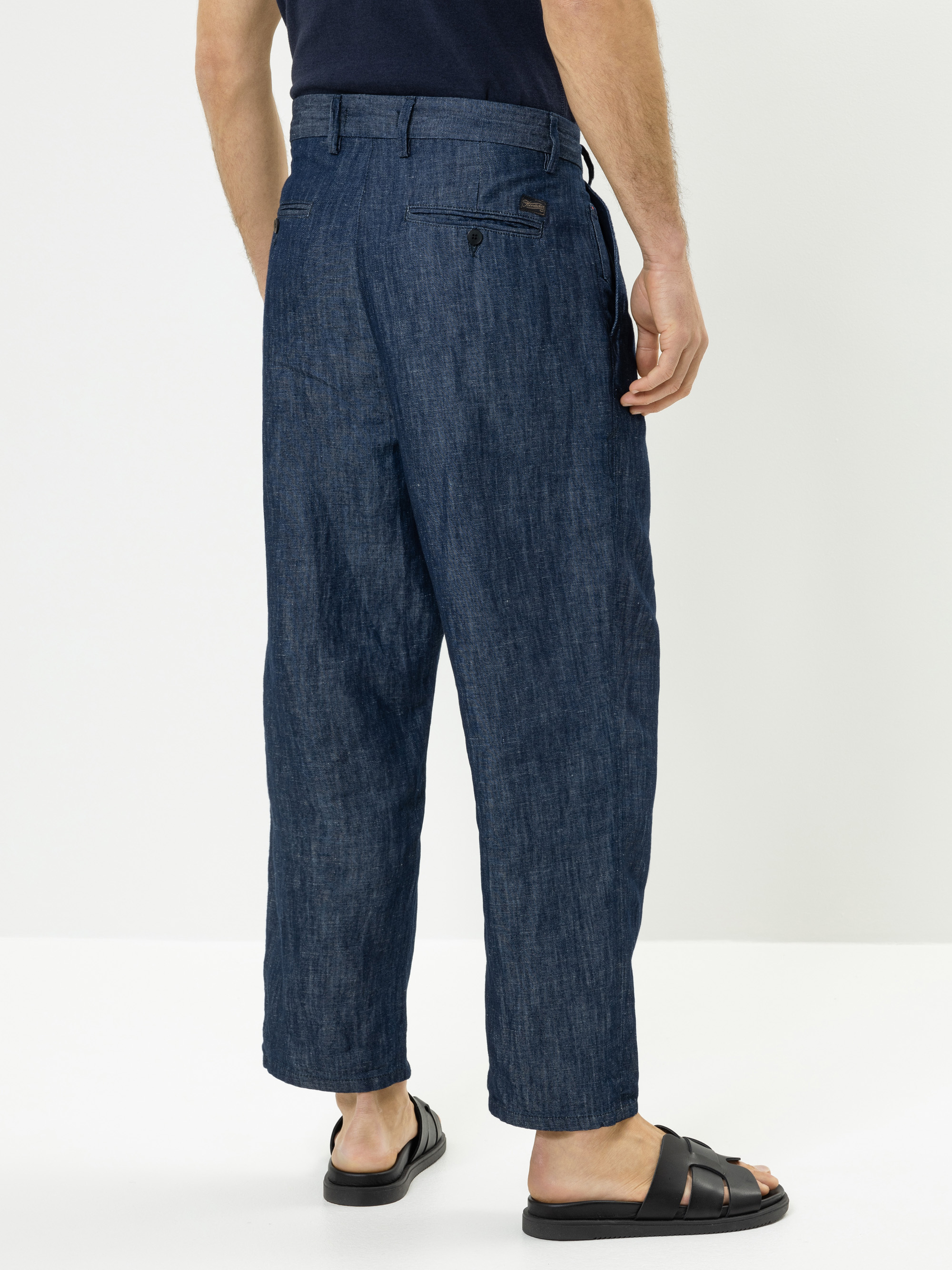 Eine Person, die mit dem Rücken zugewandt steht und eine locker sitzende blaue Jeanshose, ein dunkles Hemd und schwarze Sandalen trägt. Die Hose hat sichtbare Taschen und Gürtelschlaufen. Der Hintergrund ist einfarbig und weiß.