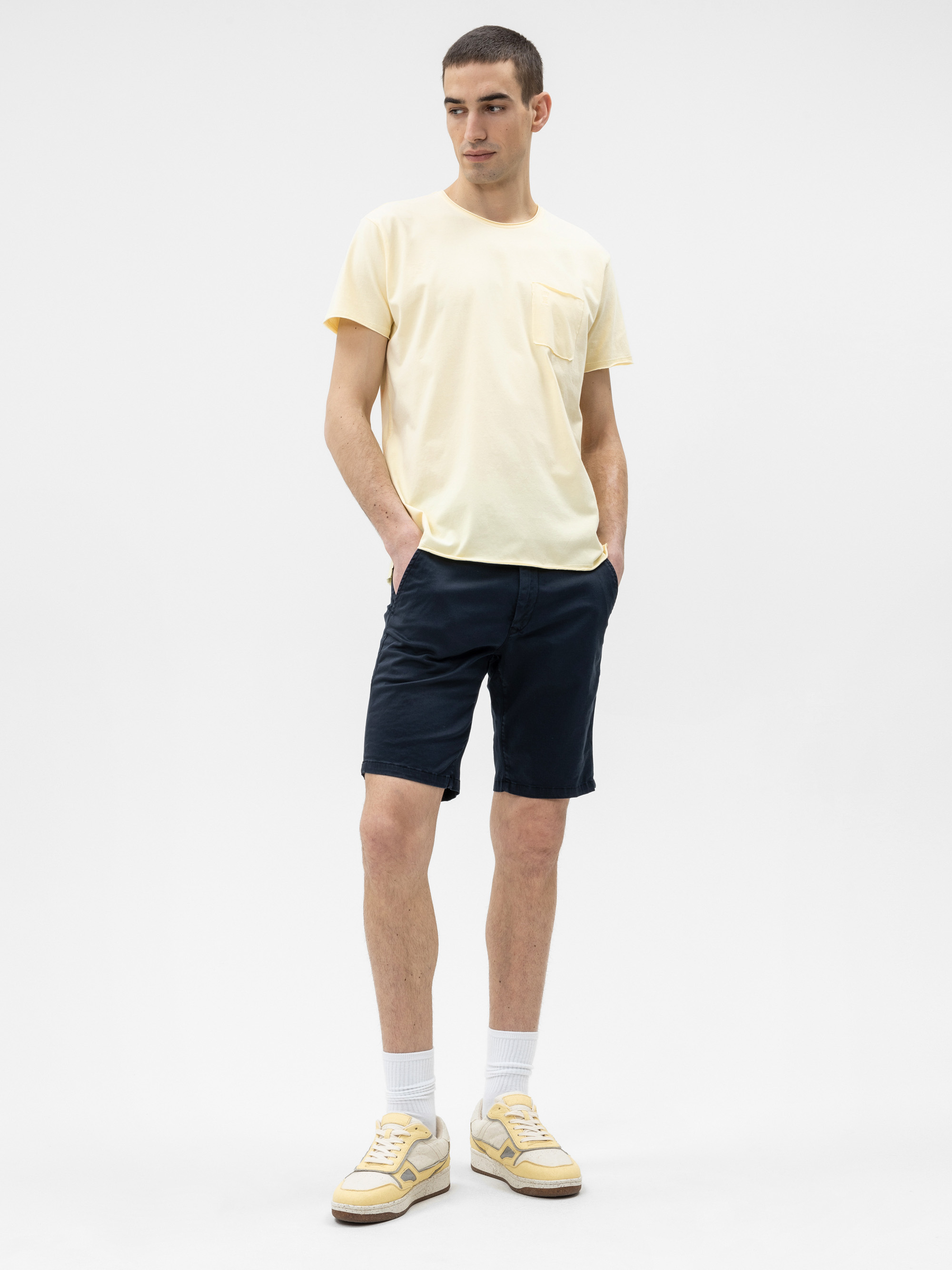 Ein Mann steht vor einem weißen Hintergrund und trägt ein blassgelbes T-Shirt, dunkle marineblaue Shorts, weiße Socken und gelb-weiße Turnschuhe. Seine Hände stecken in den Hosentaschen, und er schaut leicht zur Seite.
