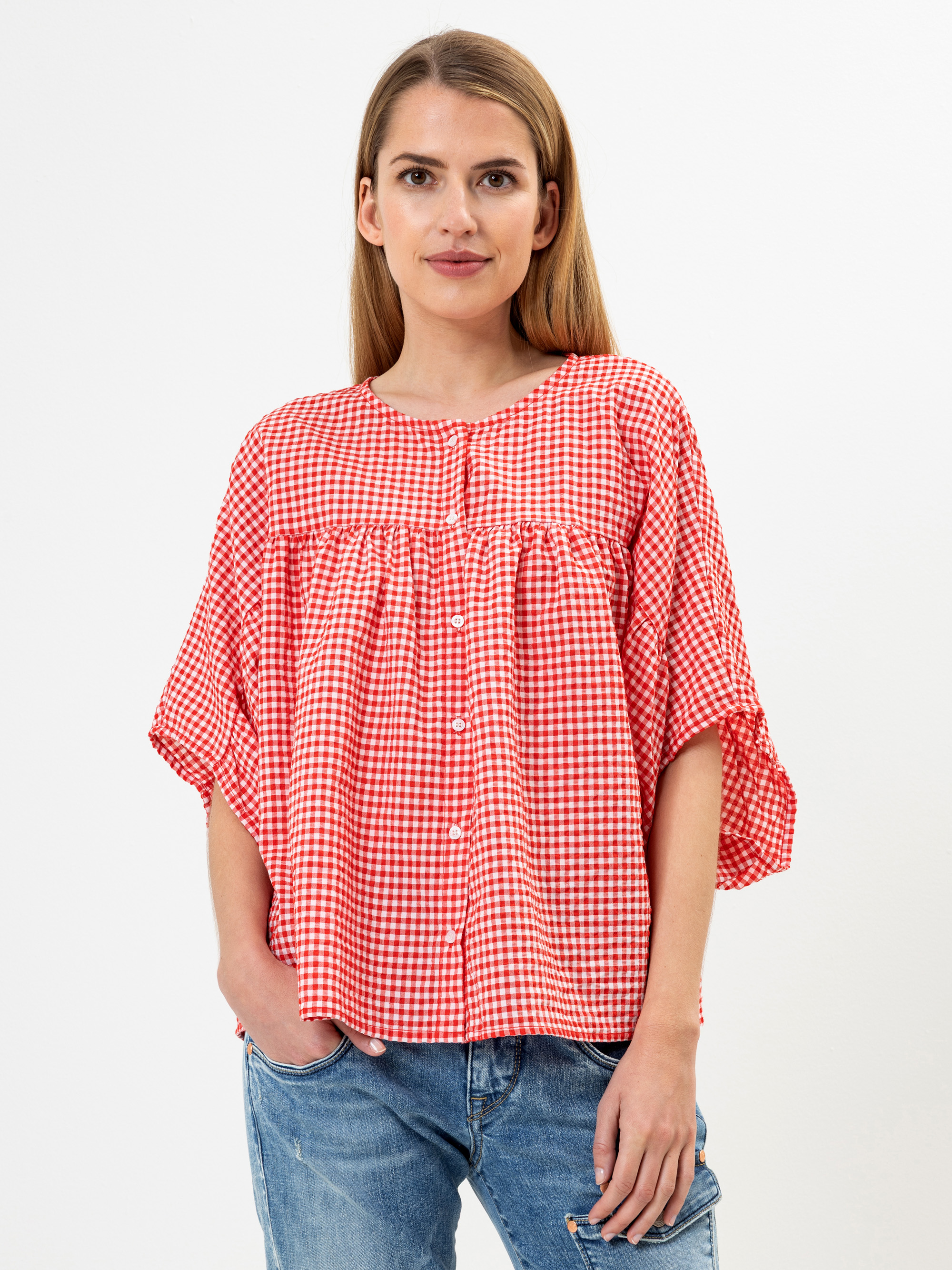Eine Frau steht vor einem weißen Hintergrund und trägt eine locker sitzende, rot-weiß karierte Button-up-Bluse mit kurzen Ärmeln und blaue Jeans. Sie hat langes, glattes Haar und einen neutralen Gesichtsausdruck.