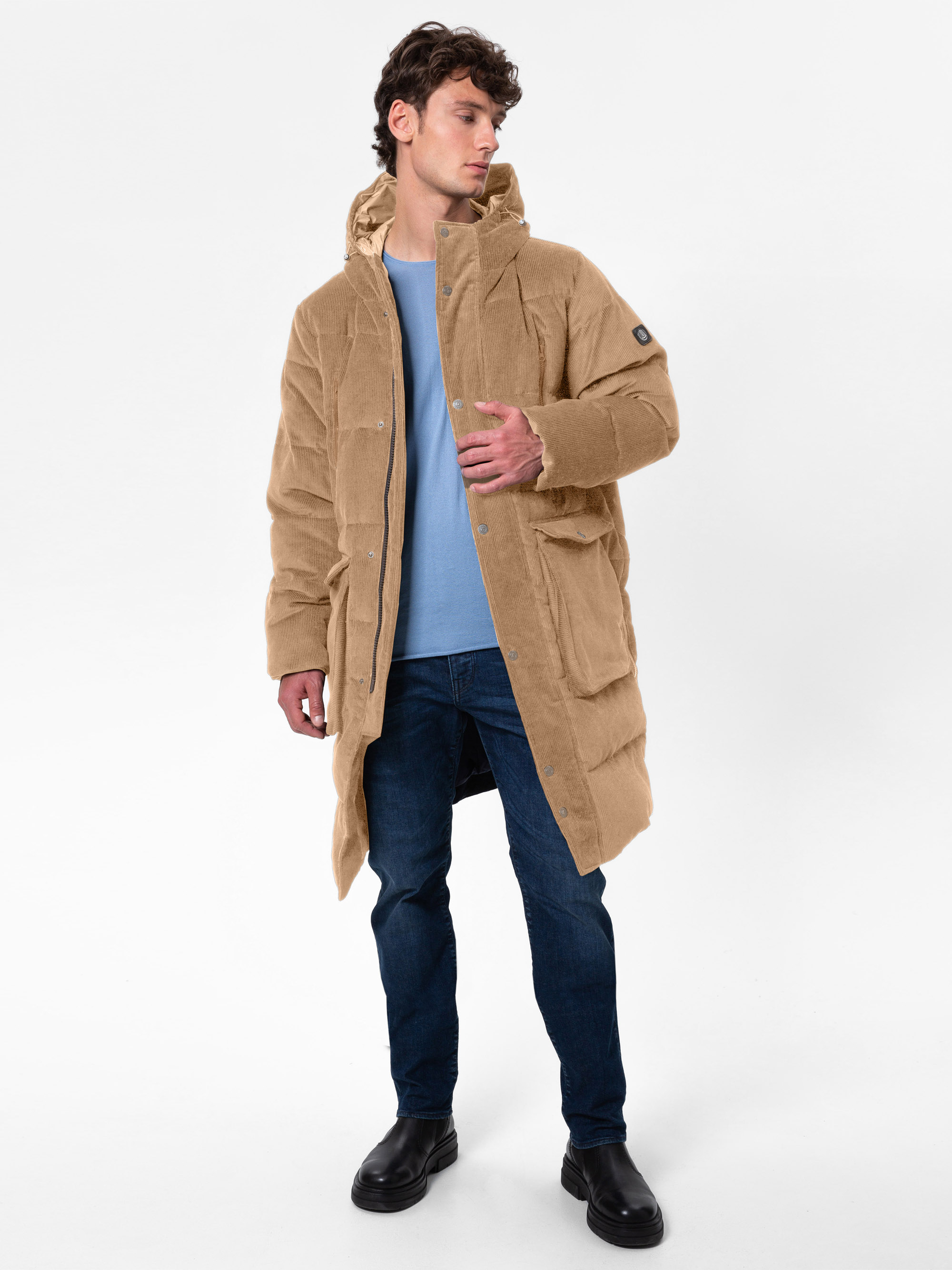 Ein Mann steht in neutraler Pose und trägt einen langen hellbraunen Kordmantel, ein hellblaues Hemd, dunkelblaue Jeans und schwarze Stiefel vor einem schlichten weißen Hintergrund.