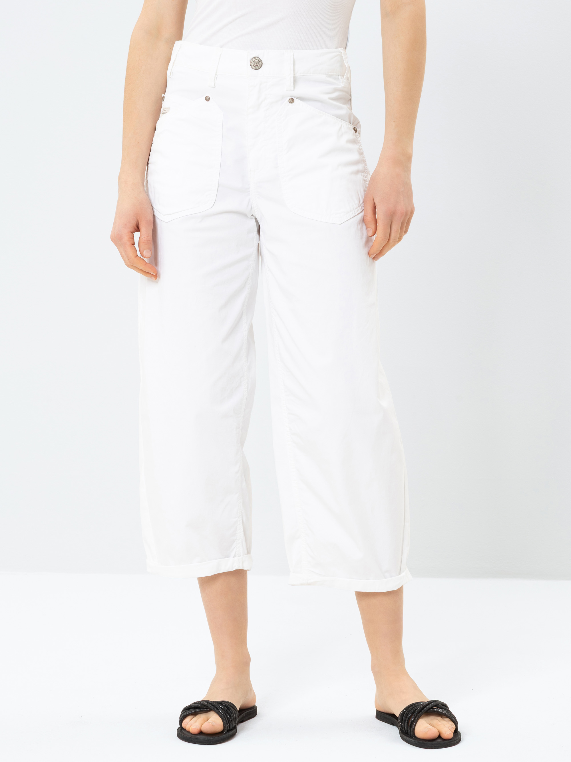 Eine Person, die eine weiße Hose mit hoher Taille und weitem Bein mit großen Taschen auf der Vorderseite und ein weißes Oberteil trägt, gepaart mit schwarzen Slippern, steht vor einem einfarbig weißen Hintergrund.