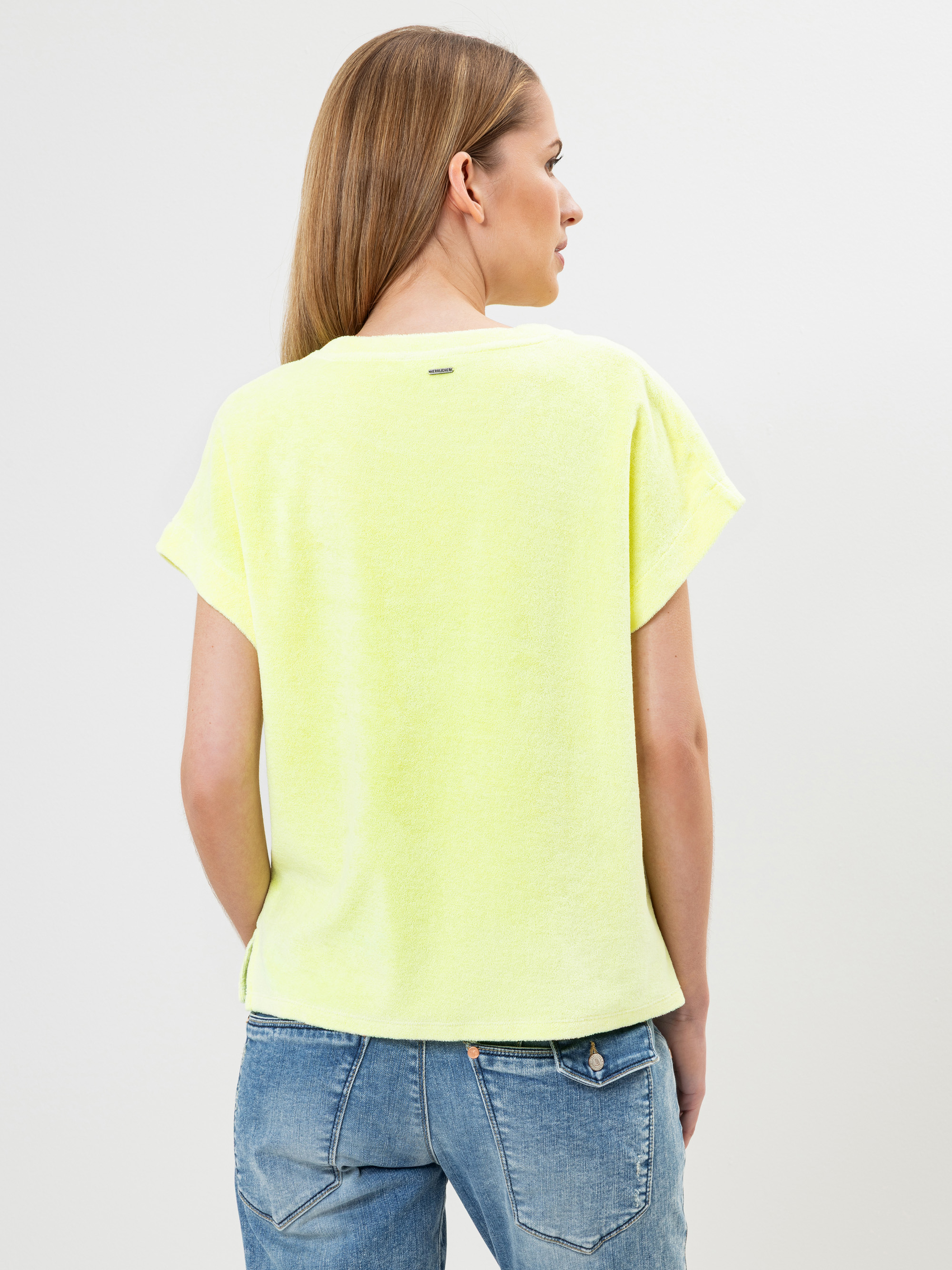 Eine Frau mit glattem, hellbraunem Haar wird von hinten gezeigt. Sie trägt ein locker sitzendes, kurzärmeliges, hellgelbes Oberteil und blaue Jeans vor einem schlichten weißen Hintergrund.