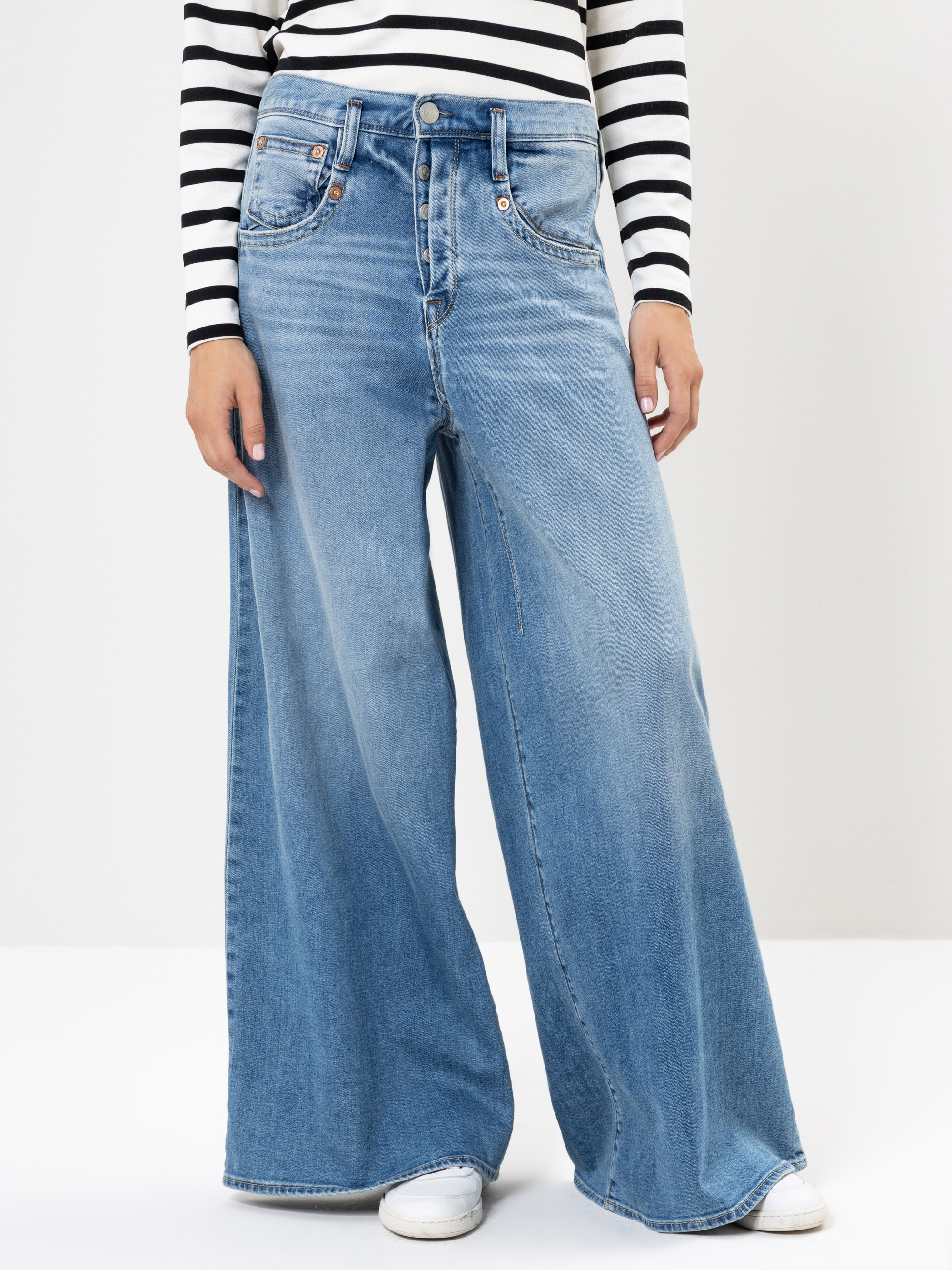 Eine Person, die ein schwarz-weiß gestreiftes langärmeliges Hemd, hellblaue Jeans mit weitem Bein und weiße Turnschuhe trägt, steht vor einem schlichten weißen Hintergrund.