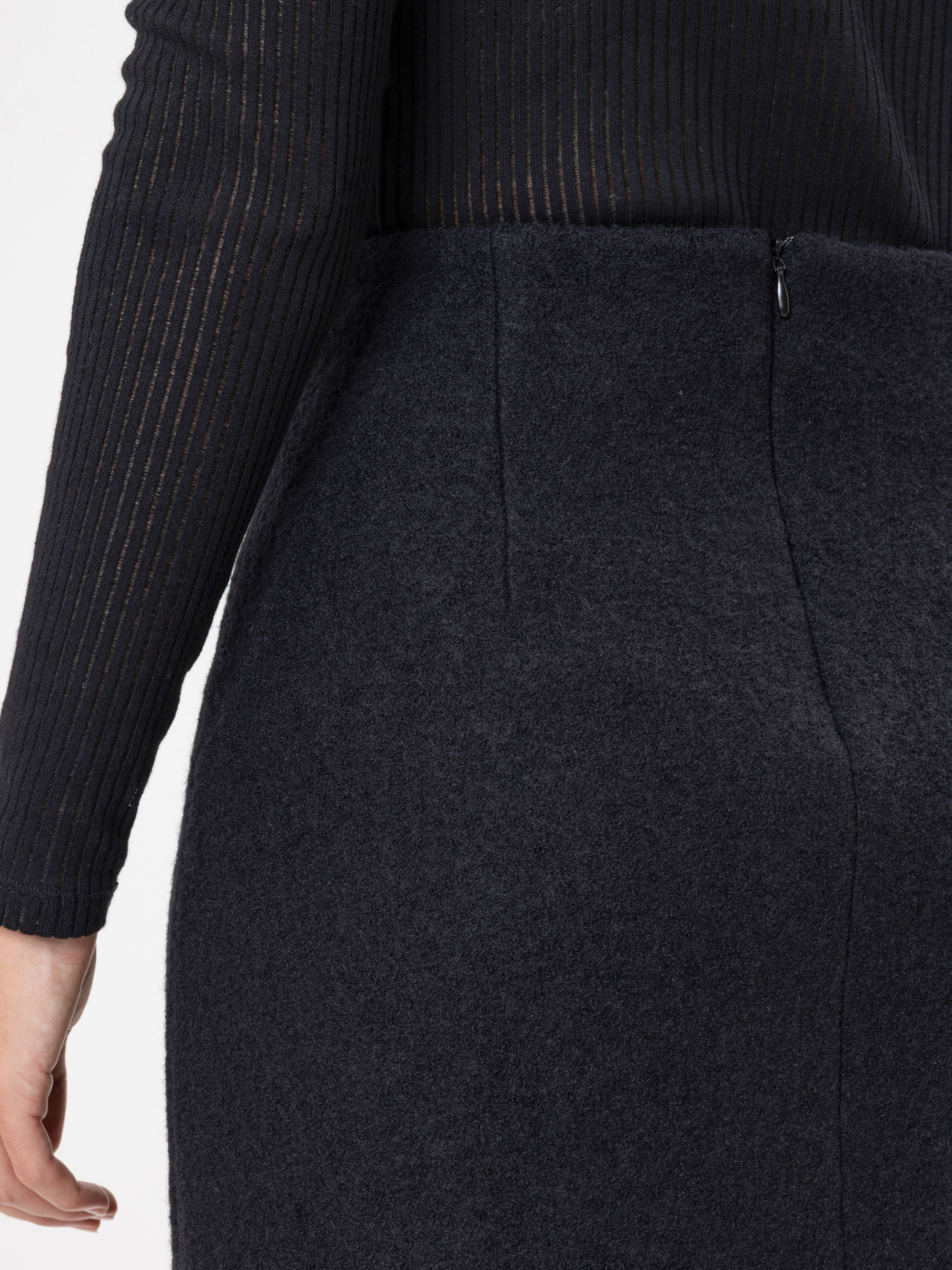 Nahaufnahme einer Person, die ein geripptes schwarzes langärmeliges Oberteil und einen hochtaillierten, dunkelgrauen Wollrock mit sichtbarem Seitenreißverschluss trägt. Nur der untere Teil des Oberkörpers und ein Teil eines Arms sind sichtbar.