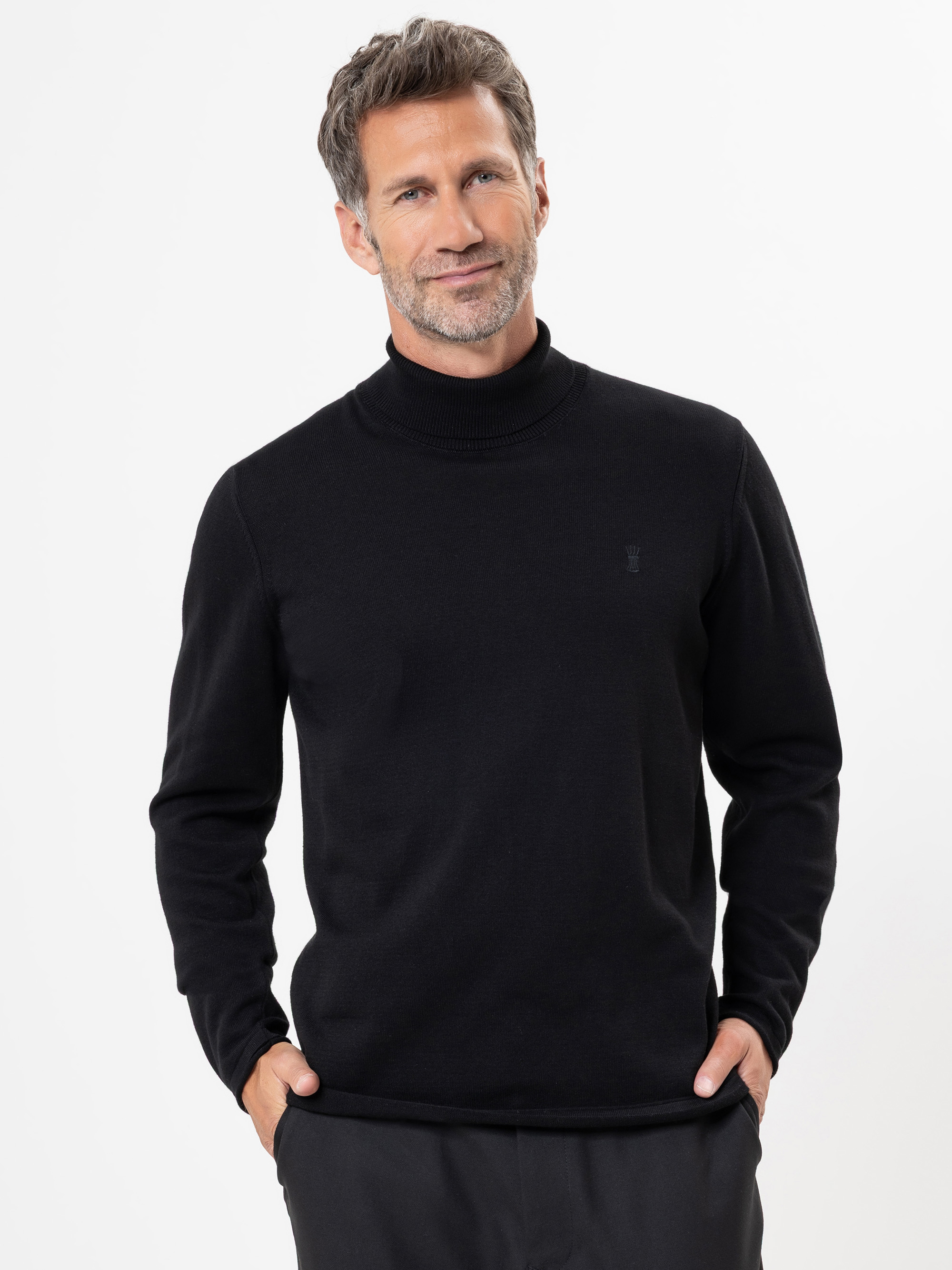 Ein Mann mit kurzem, gräulichem Haar und gestutztem Bart trägt einen schwarzen Rollkragenpullover und eine dunkle Hose. Er steht vor einem schlichten weißen Hintergrund, die Hände in den Taschen und lächelt leicht.