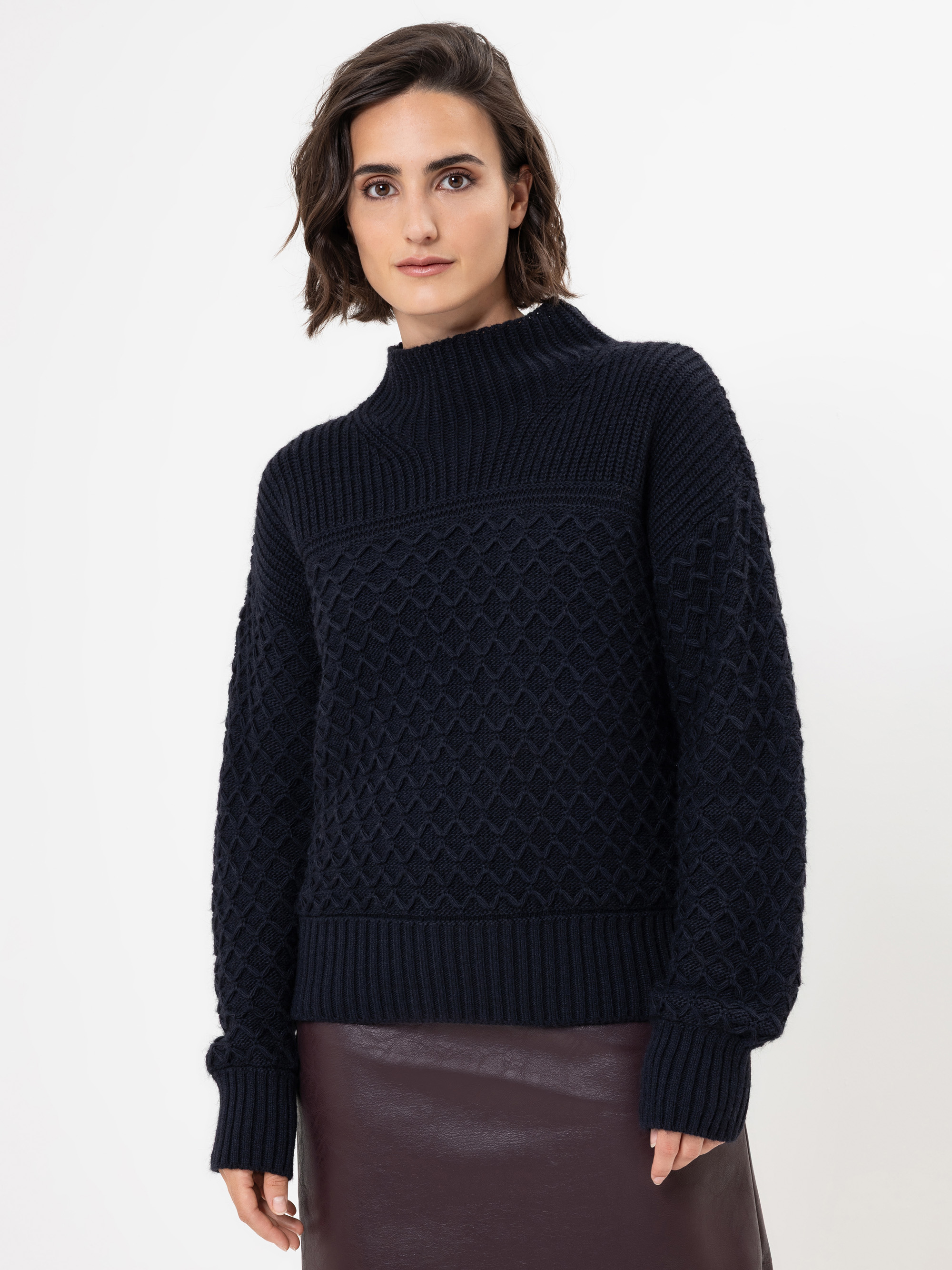 Eine Frau mit kurzen braunen Haaren, die einen strukturierten dunkelnavyfarbenen Pullover und einen dunkelbraunen Lederrock trägt, steht vor einem schlichten weißen Hintergrund und schaut mit neutralem Blick in die Kamera.