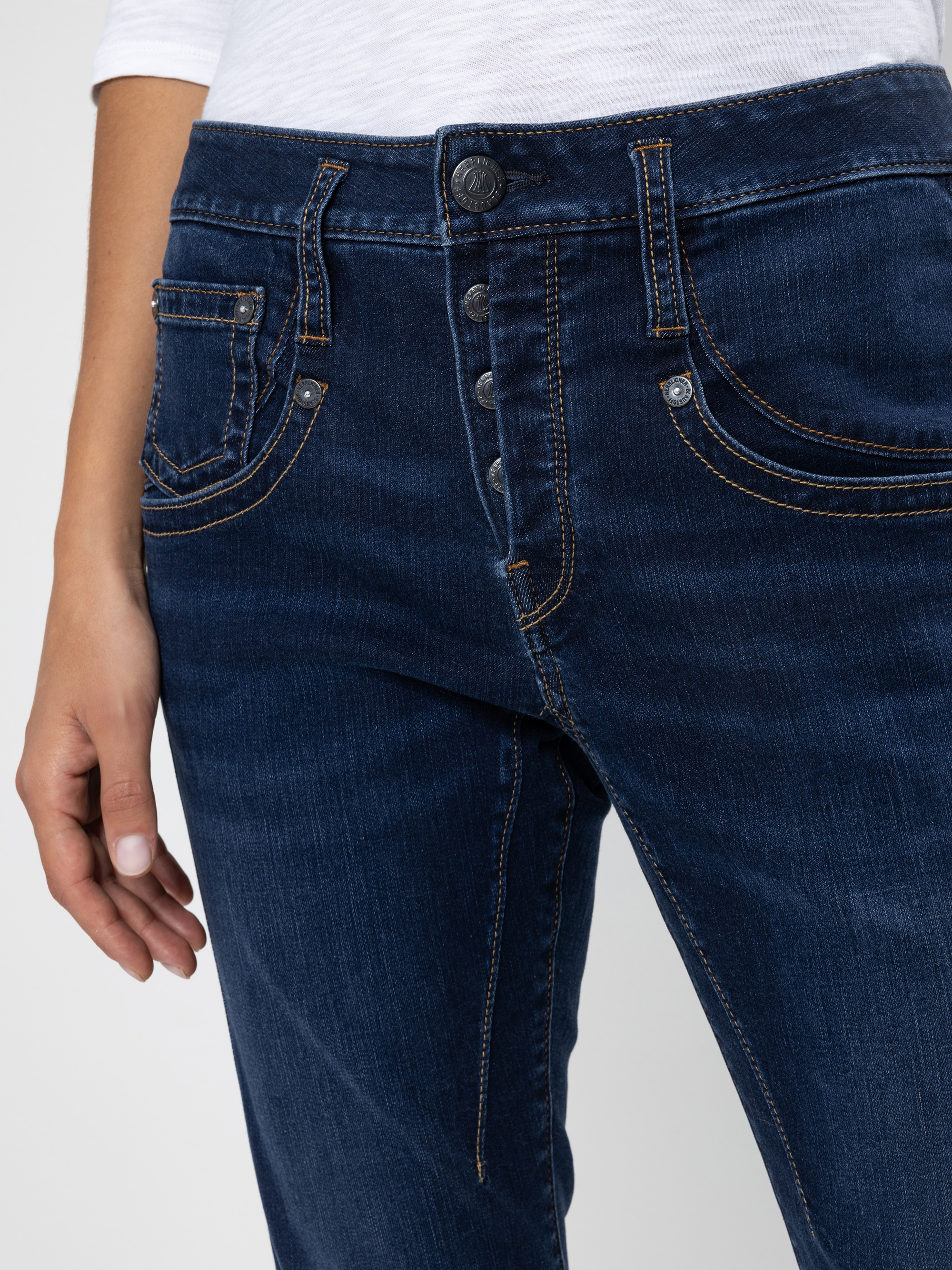 Eine Person trägt eine dunkelblaue Jeans mit Kontrastnähten und ein weißes Hemd. Der Schwerpunkt liegt auf den Vordertaschen der Jeans und dem Hosenschlitz mit Knopf, wobei die linke Hand der Person an der Seite ruht.