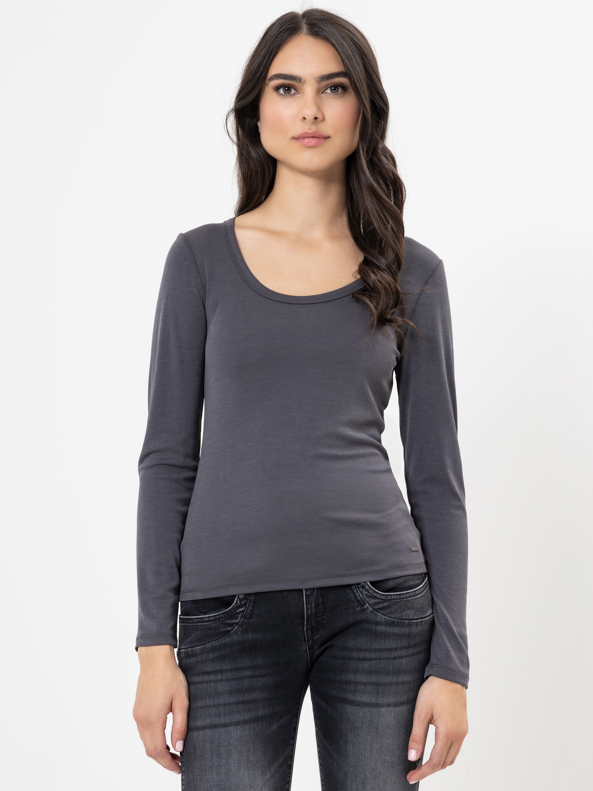 Eine Frau mit langen dunklen Haaren, die ein tailliertes, langärmeliges, anthrazitfarbenes Oberteil und dunkle Jeans trägt, steht vor einem schlichten weißen Hintergrund und schaut mit neutralem Blick geradeaus.