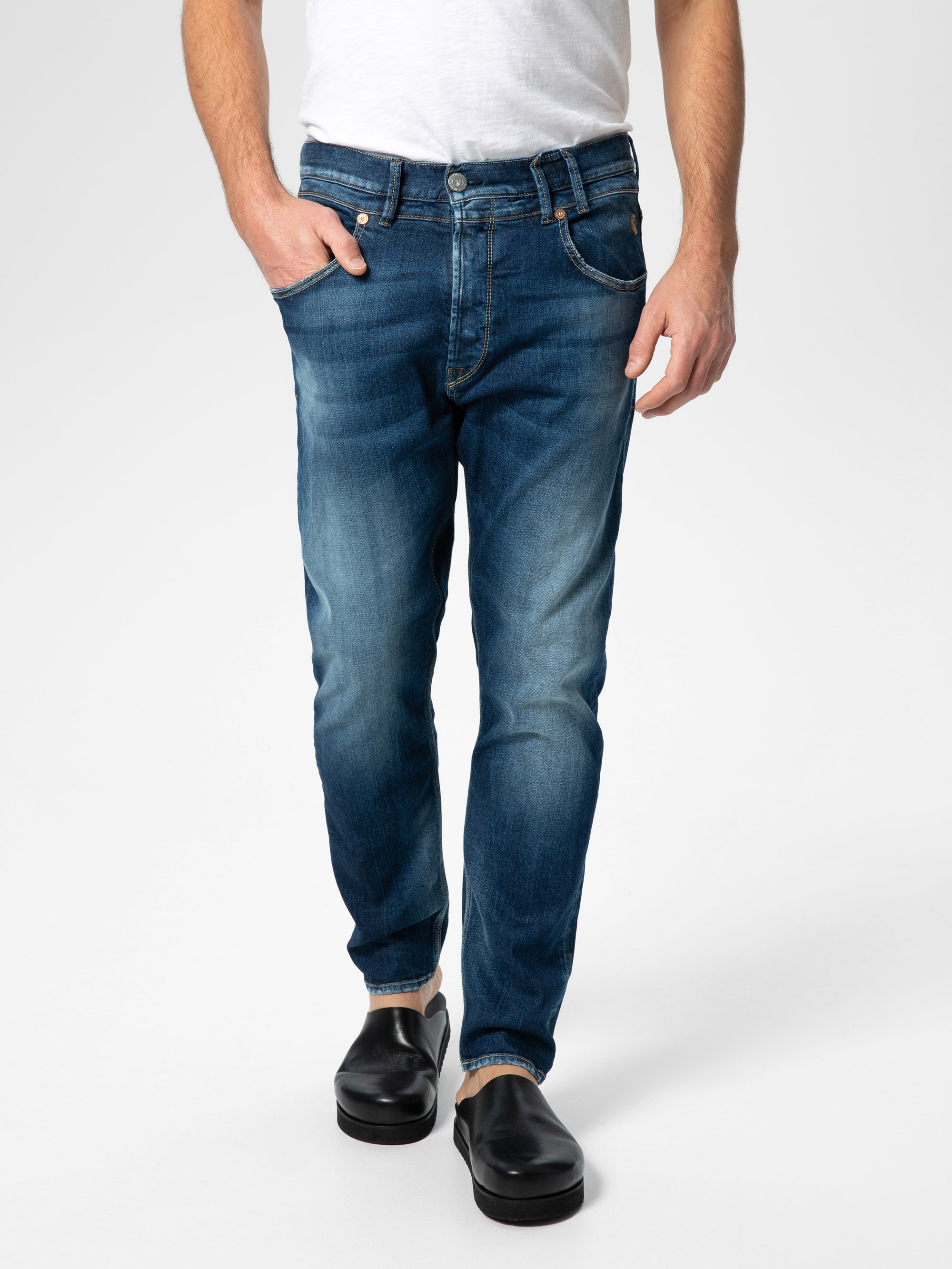 Eine Person, die ein weißes T-Shirt, blaue Jeans mit verblasster Waschung und schwarze Slipper trägt, steht vor einem schlichten hellgrauen Hintergrund. Der Oberkörper ist teilweise außerhalb des Rahmens.