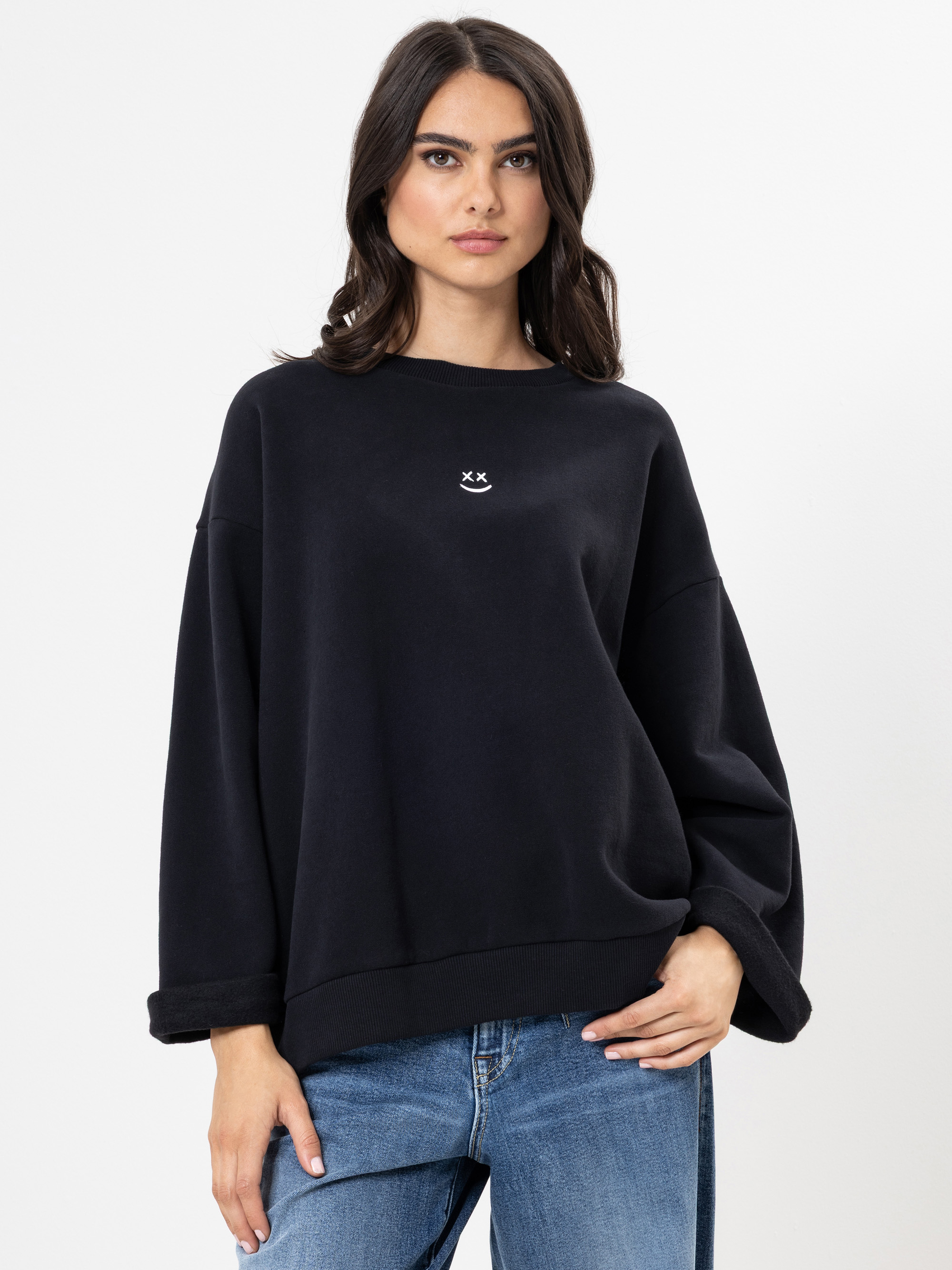Eine Frau mit langen dunklen Haaren trägt ein locker sitzendes schwarzes Sweatshirt mit einer kleinen weißen Smiley-Grafik auf der Brust und blaue Jeans und steht vor einem schlichten weißen Hintergrund.