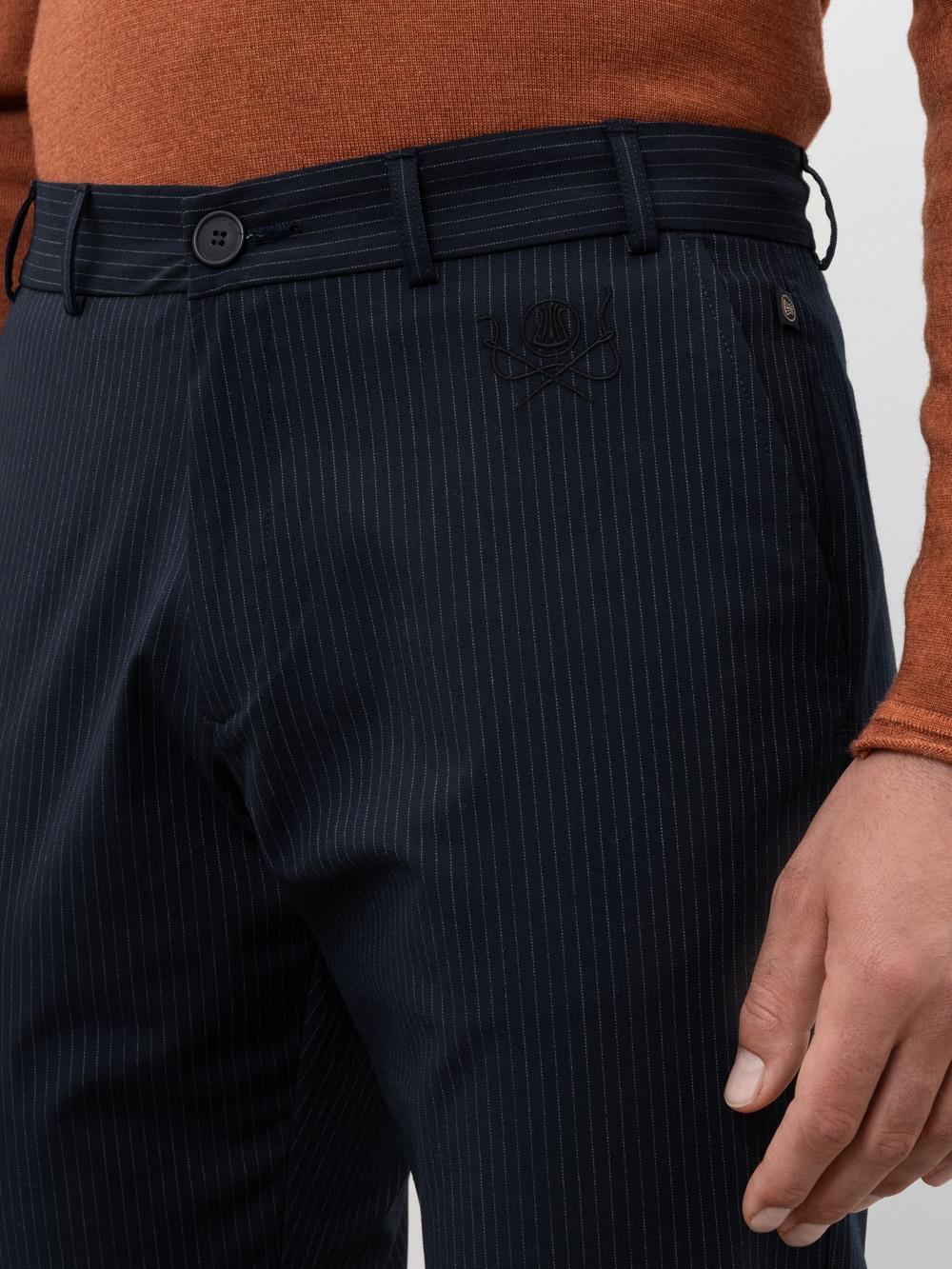 Eine Person, die eine marineblaue Nadelstreifenhose mit einem dezent aufgestickten Totenkopf-Logo in der Nähe der Tasche trägt, gepaart mit einem rostfarbenen langärmeligen Oberteil. Nur der untere Teil des Oberkörpers und die Hand sind sichtbar.