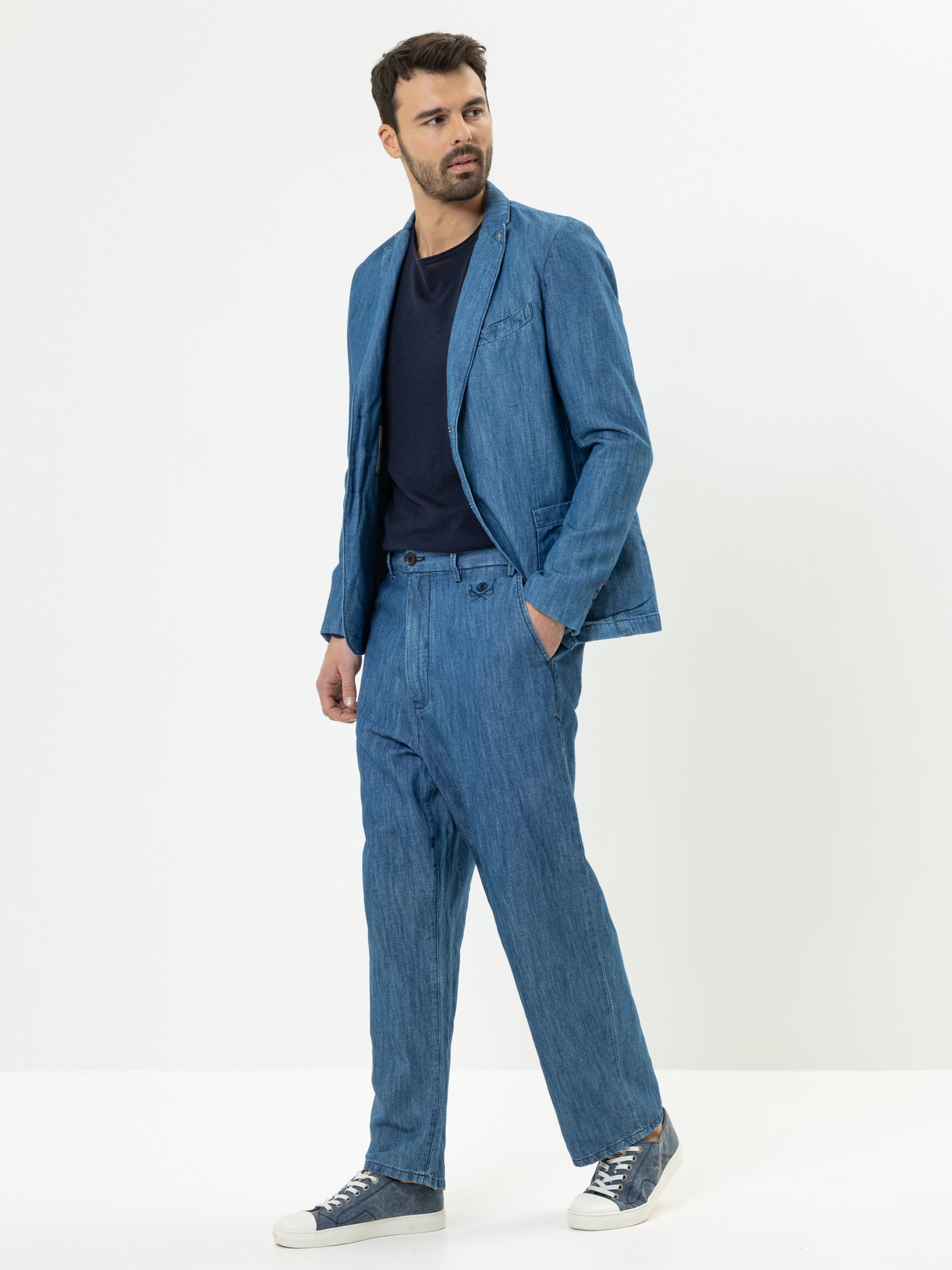 Ein Mann, der einen lässigen blauen Jeansanzug mit einem schwarzen T-Shirt und blauen Turnschuhen trägt, steht vor einem schlichten weißen Hintergrund und schaut leicht zur Seite.