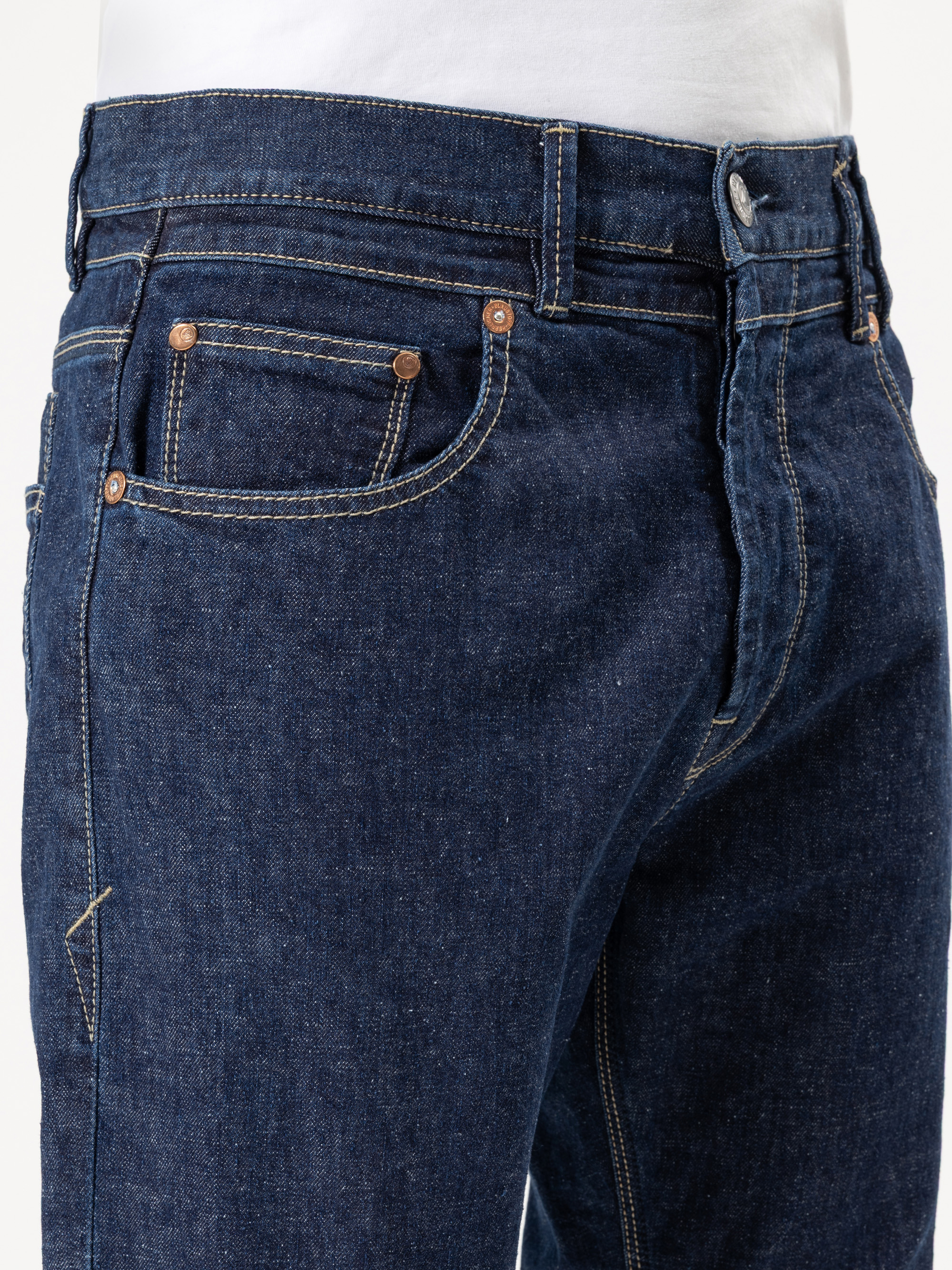 Nahaufnahme einer Person, die eine dunkelblaue Jeans und ein weißes Hemd trägt, mit Schwerpunkt auf der Taille, den Taschen und dem Oberschenkelbereich. Die Jeans weist sichtbare Nähte sowie silberne und kupferne Nieten auf.