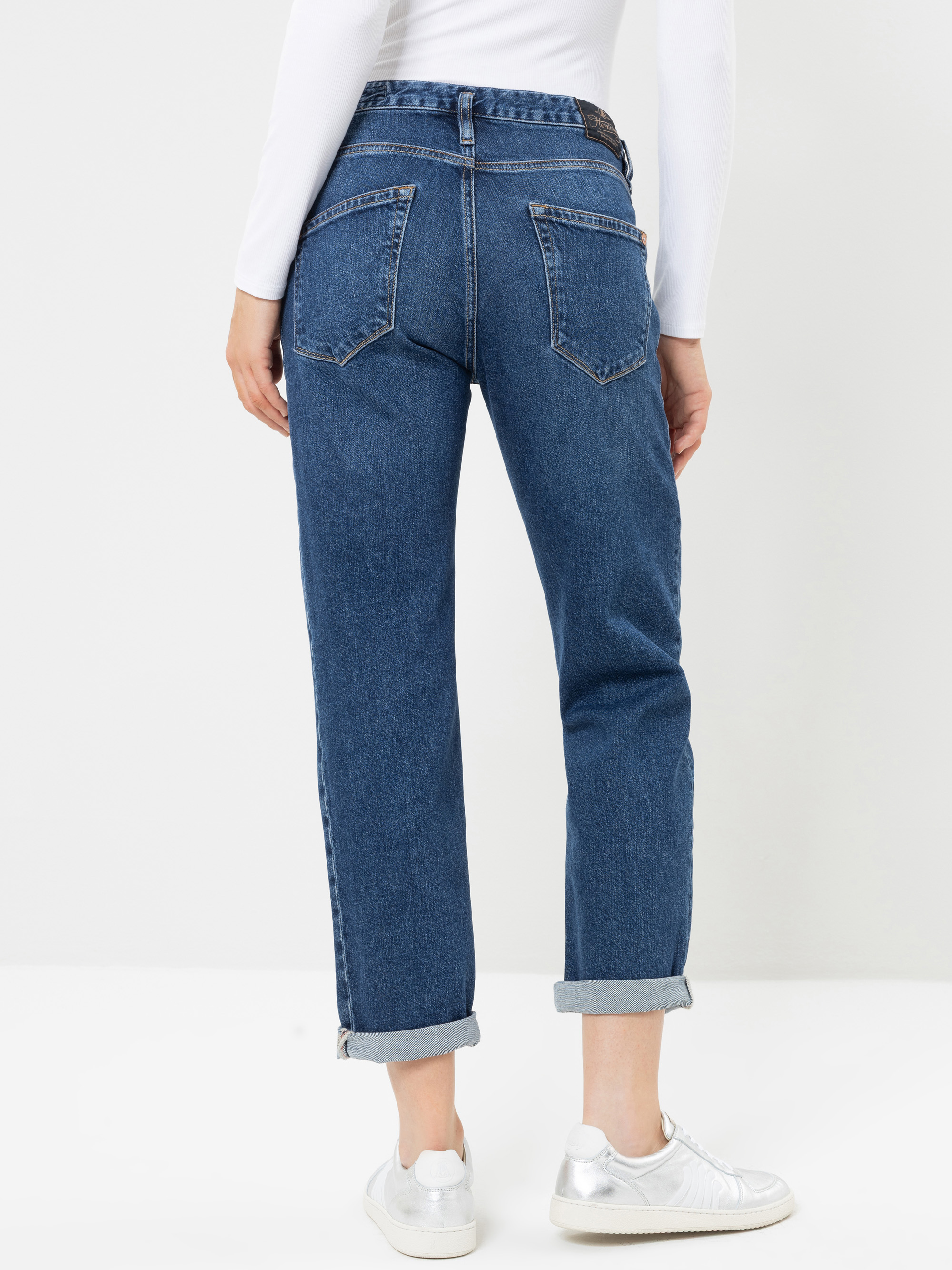 Eine Person, die eine blaue Jeans mit Umschlag und ein weißes Langarm-Oberteil trägt, von hinten gesehen. Die Jeans hat große Gesäßtaschen, und die Person steht auf einem weißen Untergrund und trägt weiße Turnschuhe.