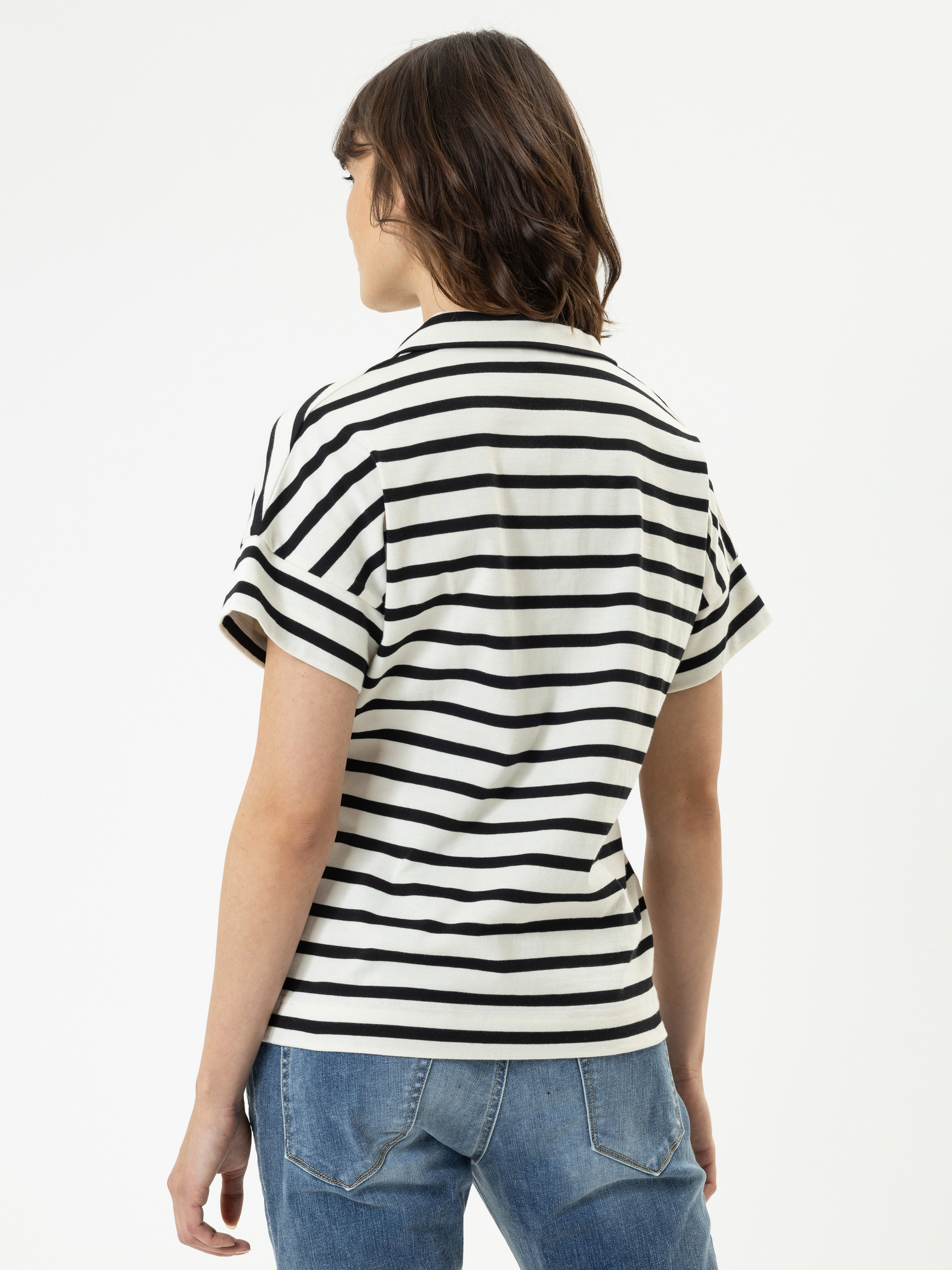 Man sieht eine Person mit schulterlangen braunen Haaren von hinten, die ein kurzärmeliges weißes T-Shirt mit schwarzen Querstreifen und eine blaue Jeans trägt und vor einem schlichten weißen Hintergrund steht.