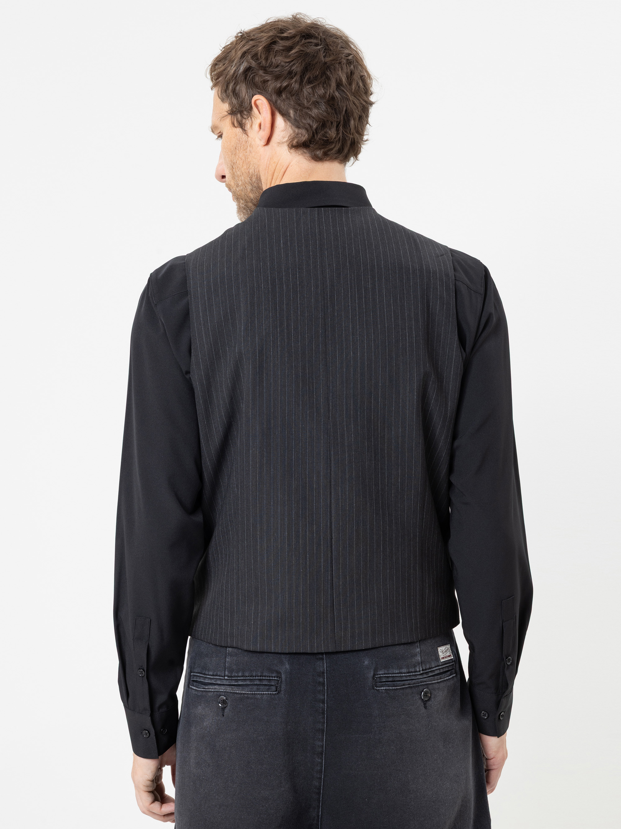 Ein Mann steht mit dem Rücken zur Kamera und trägt eine schwarze Nadelstreifenweste über einem schwarzen Hemd und einer dunklen Hose. Der Hintergrund ist schlicht weiß.