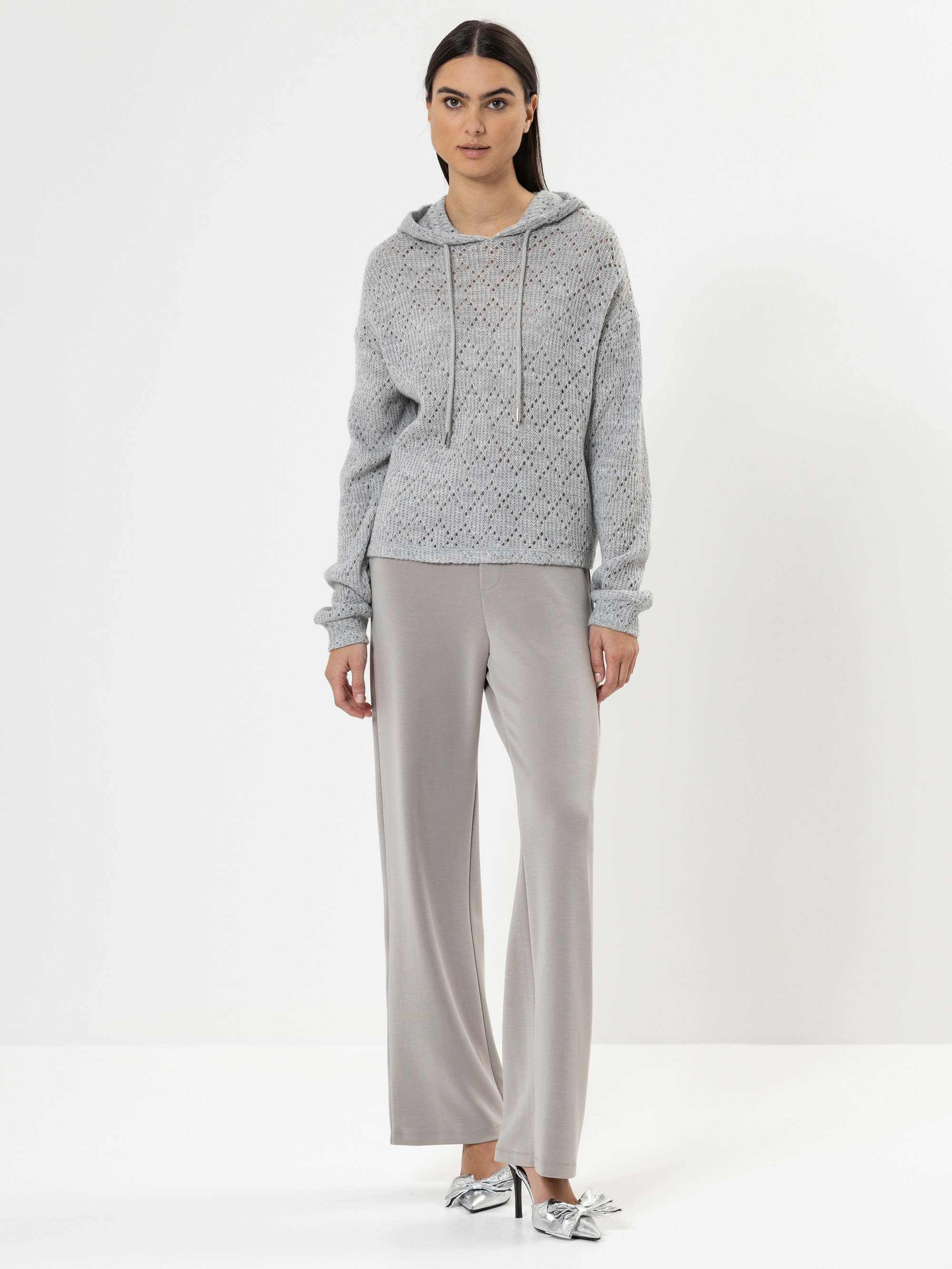 Eine Frau steht vor einem weißen Hintergrund und trägt einen hellgrauen, gestrickten Kapuzenpullover, eine weite, hellbeige Hose und mit einer silbernen Schleife verzierte Schuhe. Sie hat glattes dunkles Haar und einen neutralen Gesichtsausdruck.