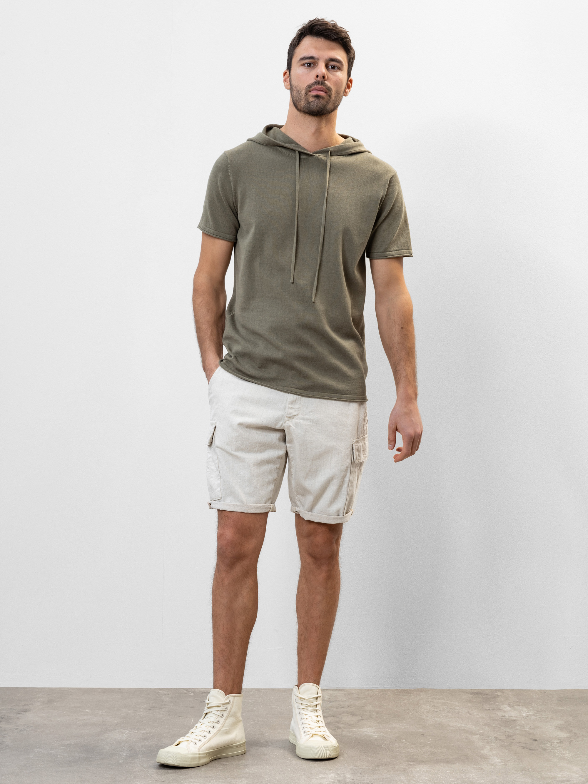 Ein Mann steht vor einer schlichten weißen Wand und trägt einen kurzärmeligen, olivgrünen Kapuzenpullover, beigefarbene Cargoshorts und cremefarbene High-Top-Turnschuhe. Er hat eine Hand in der Tasche und einen neutralen Gesichtsausdruck.