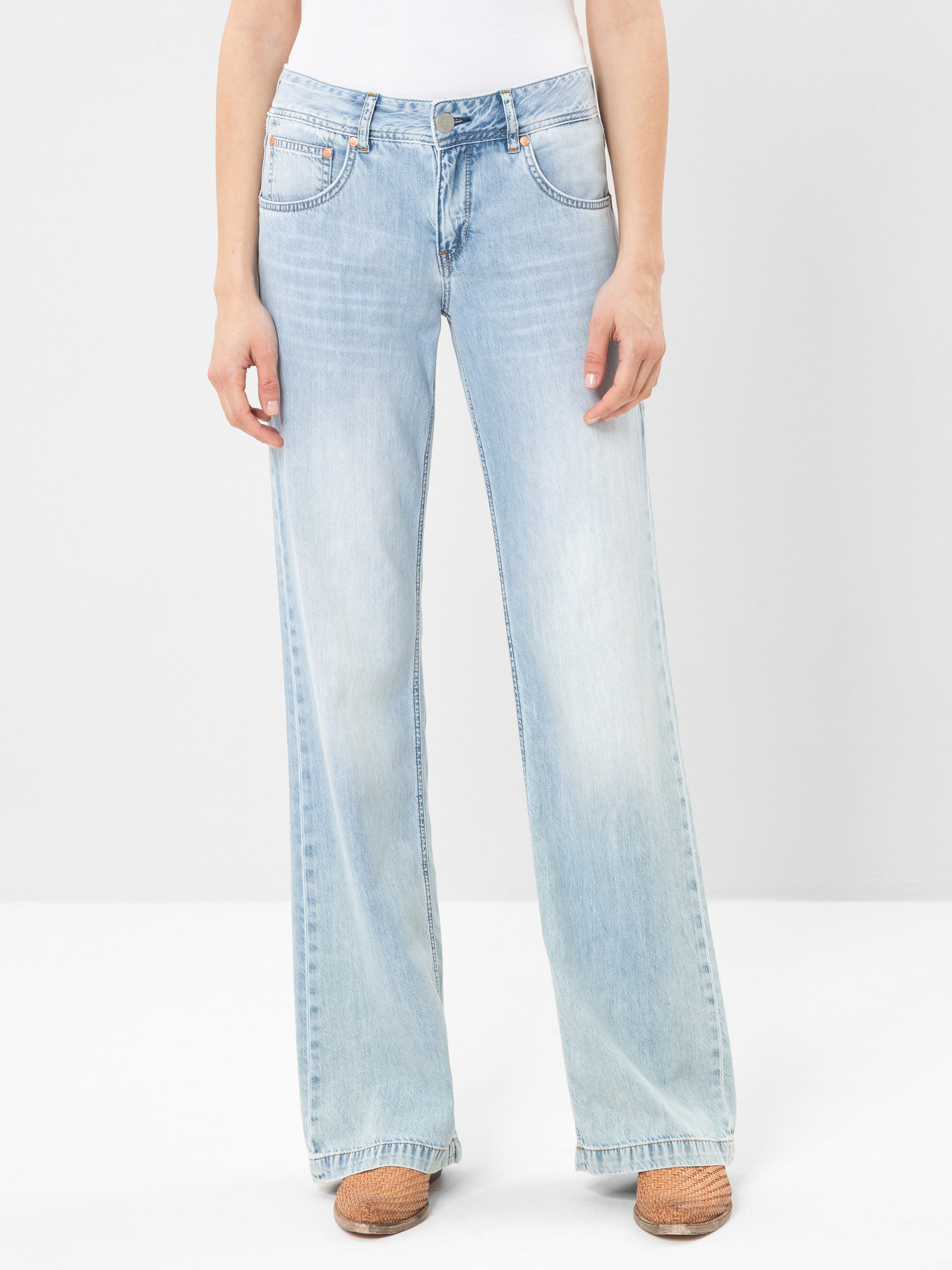 Eine Person, die hellblaue, weit geschnittene Jeans, ein weißes Oberteil und braune, gewebte Schuhe trägt, steht vor einem schlichten weißen Hintergrund. Das Bild zeigt die untere Hälfte des Körpers.