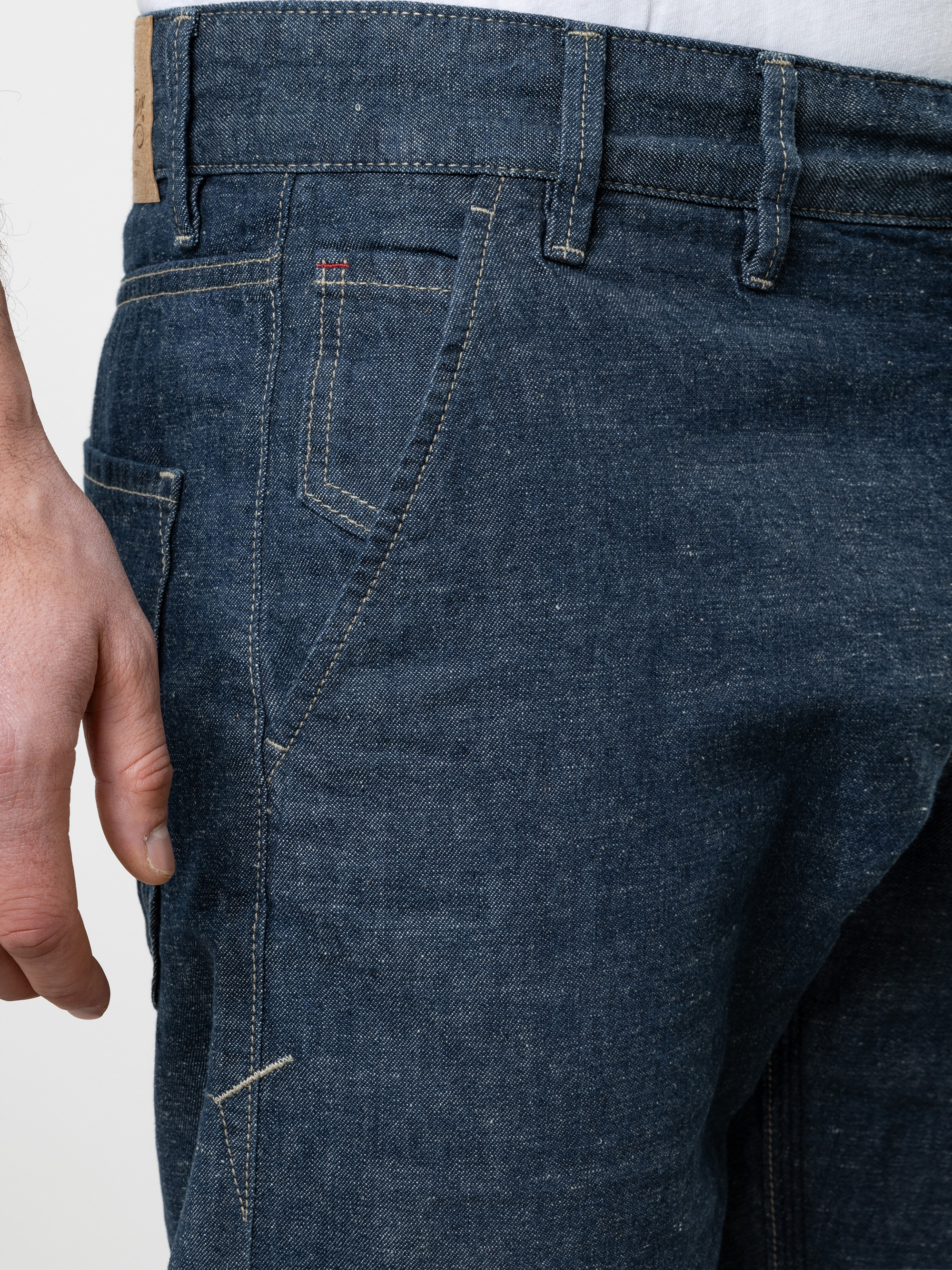 Nahaufnahme einer Person, die eine blaue Denim-Jeans trägt, mit der linken Vordertasche, der Gürtelschlaufe und einem Teil der linken Hand, die an der Seite liegt. Der Hintergrund ist einfarbig und hell.