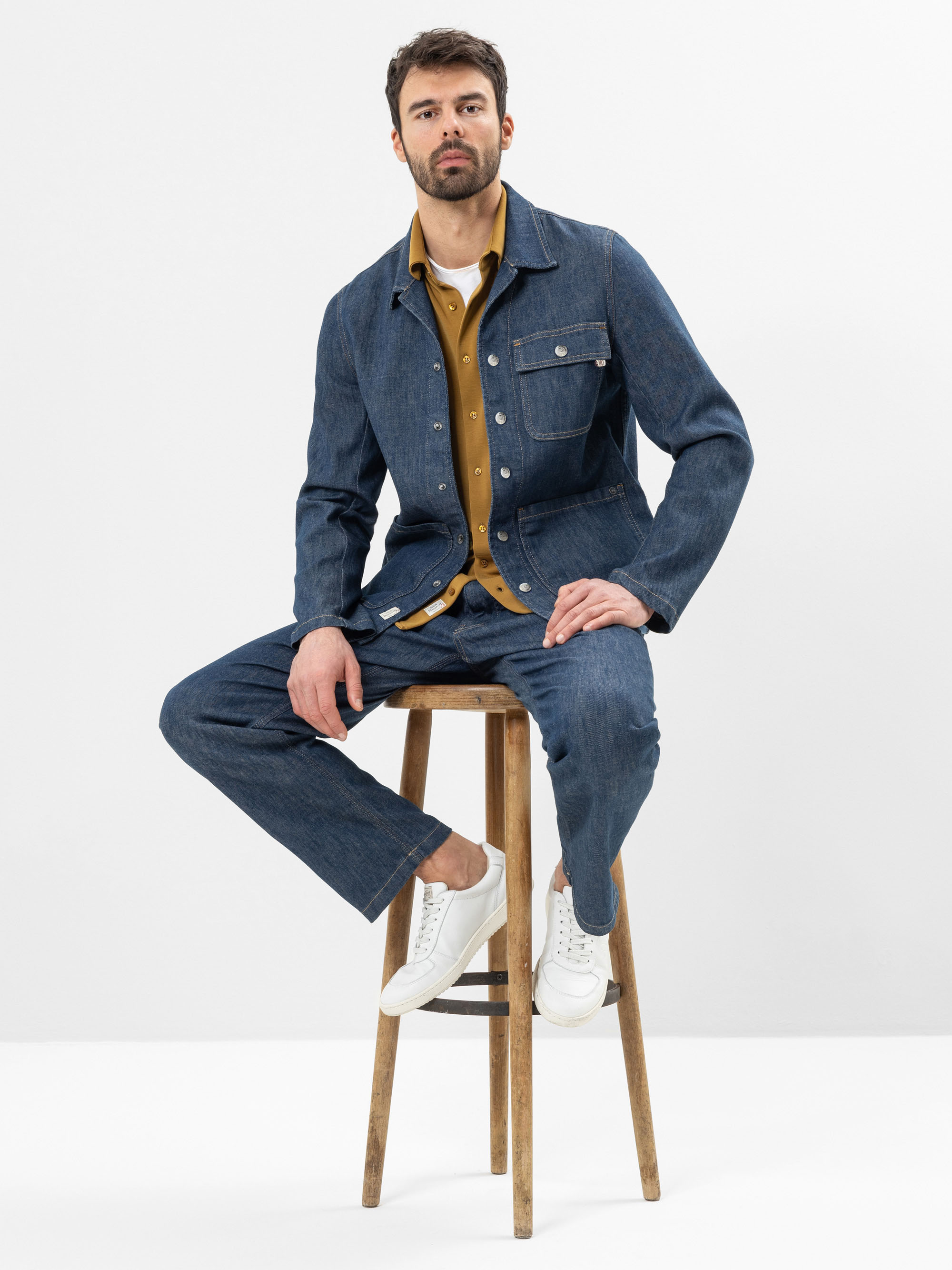Ein bärtiger Mann sitzt auf einem Holzschemel vor einem schlichten weißen Hintergrund. Er trägt eine Jeansjacke und -hose, ein senfgelbes Button-up-Hemd und weiße Turnschuhe.