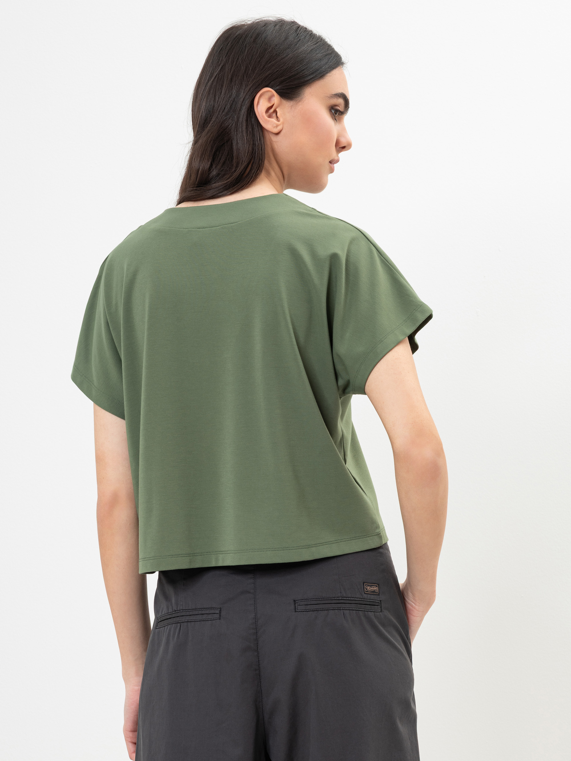 Eine Frau mit langen dunklen Haaren, die ein olivgrünes kurzärmeliges Oberteil und eine dunkelgraue Hose trägt, steht mit dem Rücken zur Kamera vor einem weißen Hintergrund.