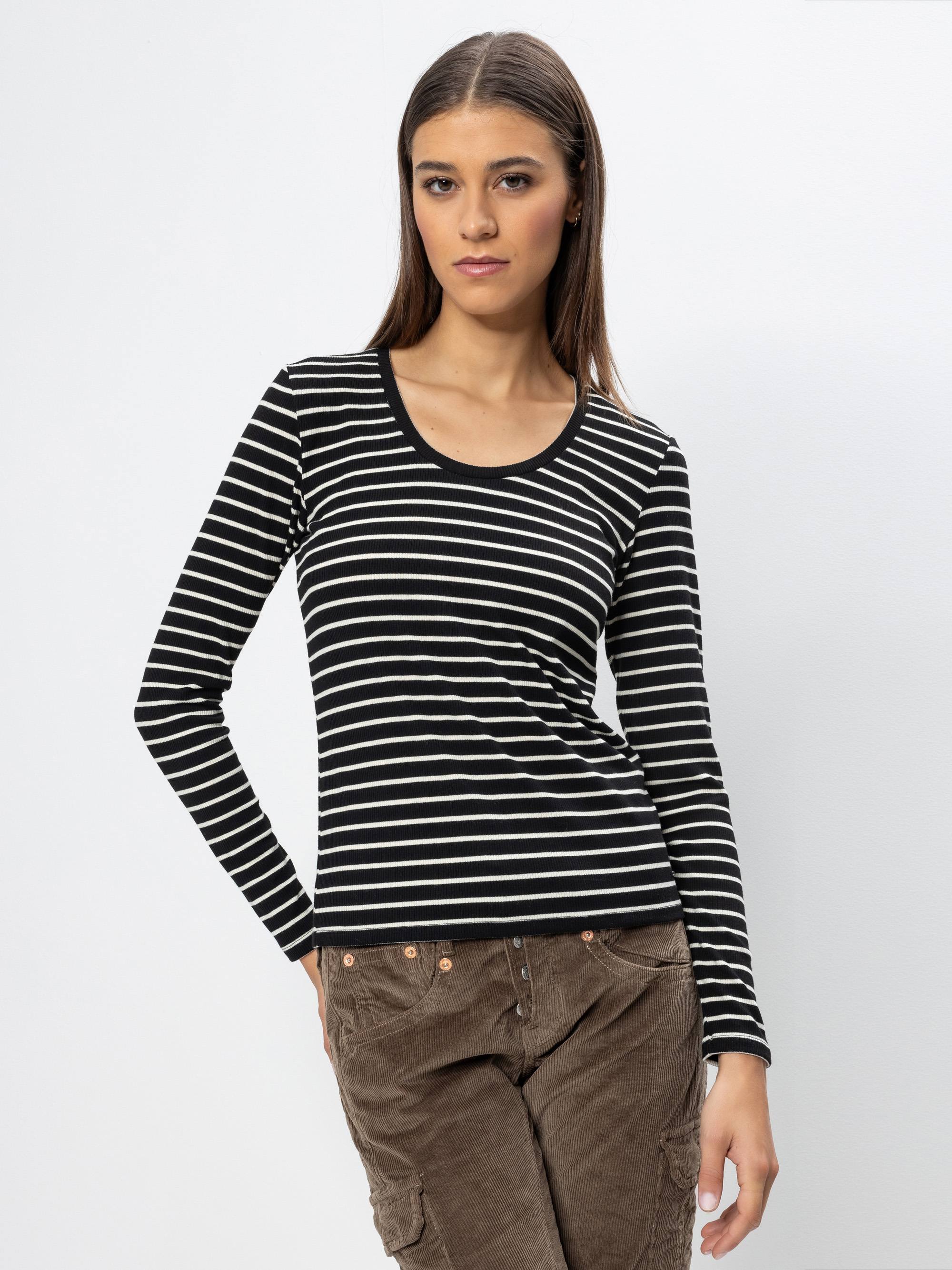 Eine Frau, die ein schwarz-weiß gestreiftes langärmeliges Oberteil und eine braune Cordhose trägt, steht vor einem schlichten weißen Hintergrund und blickt selbstbewusst in die Kamera, eine Hand in die Hüfte gestützt.