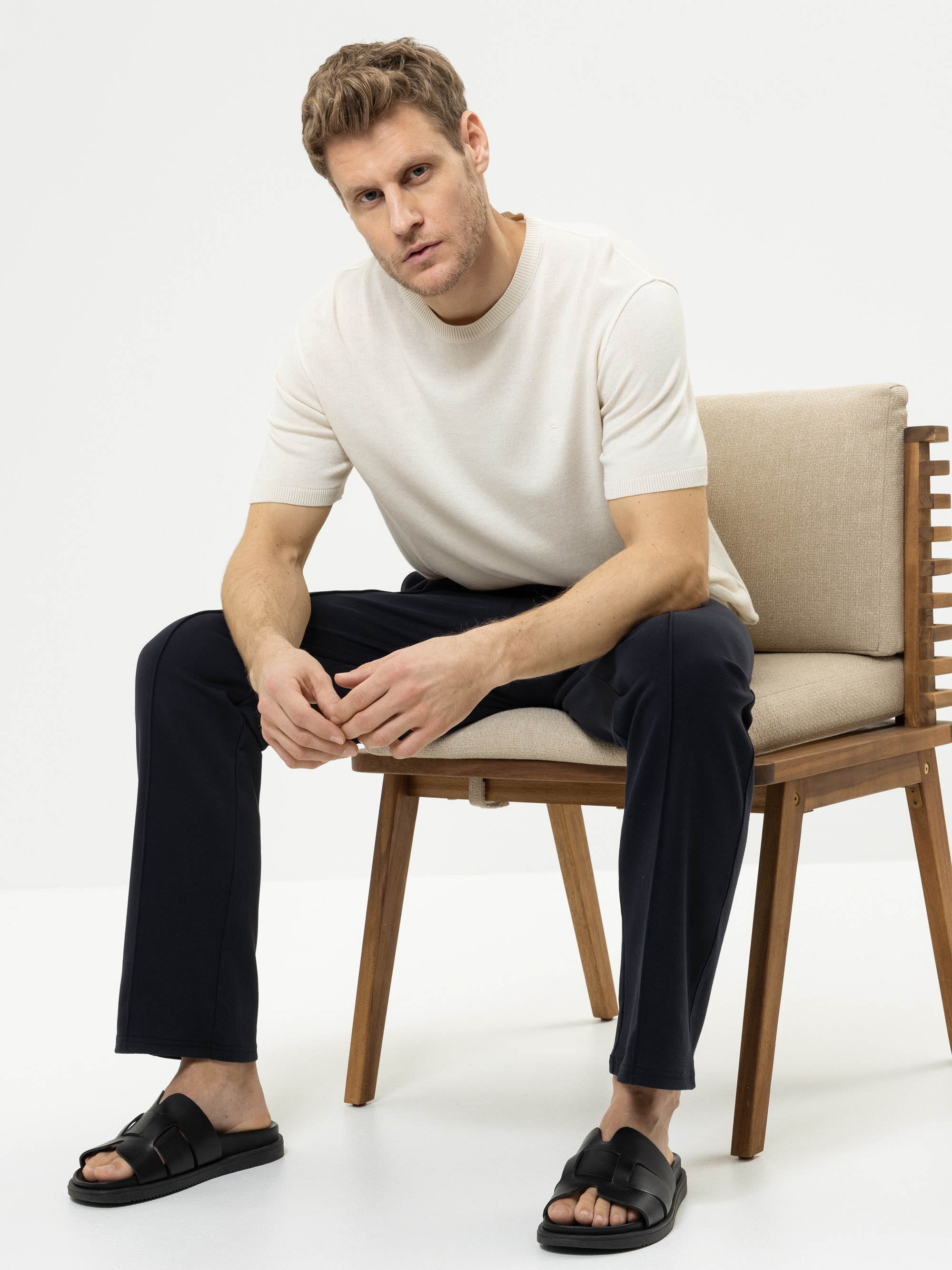 Ein Mann mit kurzen hellbraunen Haaren sitzt auf einem Holzstuhl und trägt ein cremefarbenes kurzärmeliges Hemd, eine dunkle Hose und schwarze Sandalen. Er blickt mit neutraler Miene in die Kamera vor einem schlichten weißen Hintergrund.
