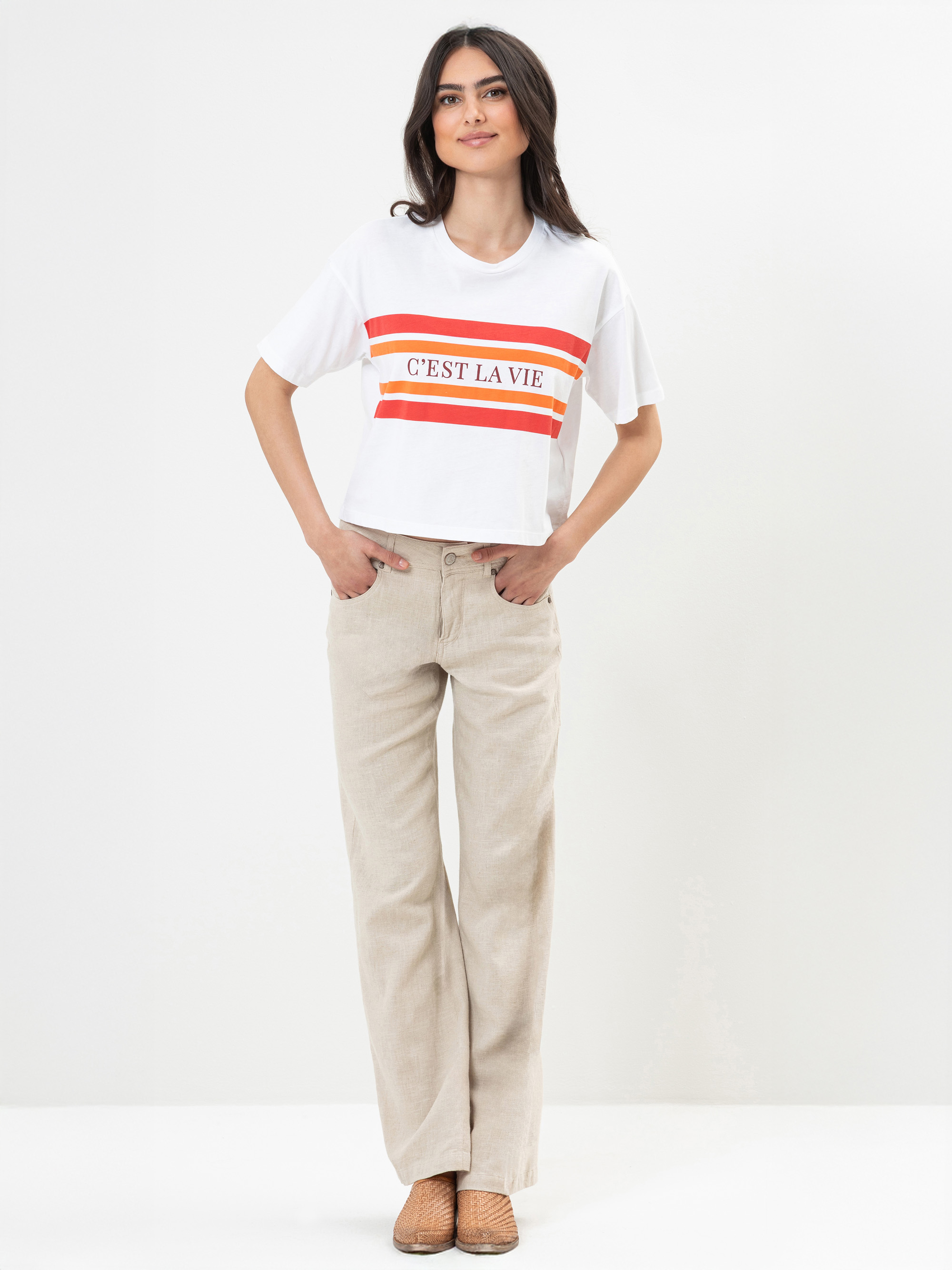 Eine Frau steht vor einem schlichten Hintergrund und trägt ein weißes T-Shirt mit roten Streifen und der Aufschrift CEST LA VIE, eine beigefarbene Hose und braune gewebte Schuhe. Sie hat lange dunkle Haare und lächelt mit den Händen in den Taschen.