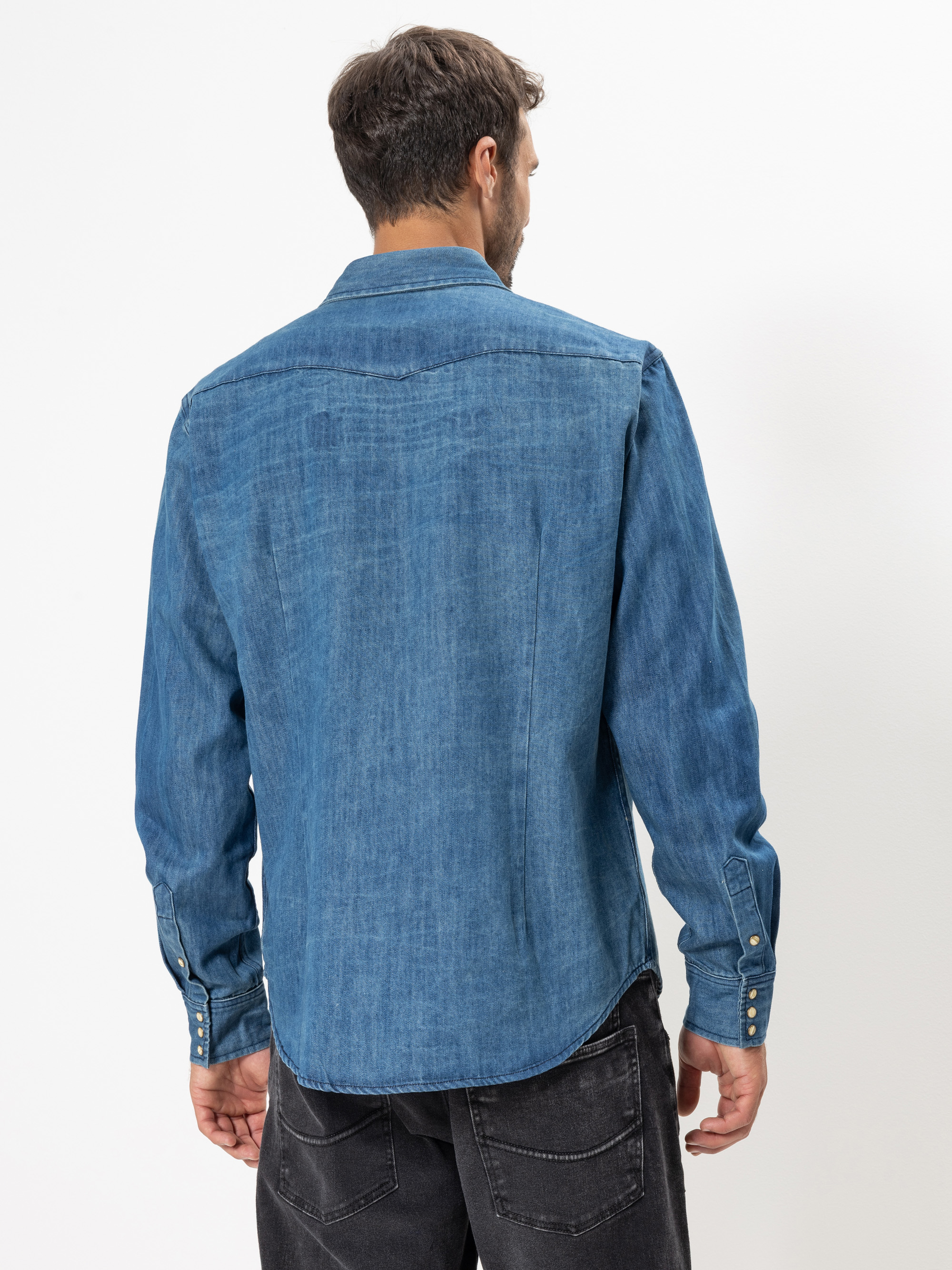 Ein Mann mit kurzen braunen Haaren blickt von sich weg, trägt ein langärmeliges blaues Jeanshemd und schwarze Jeans und steht vor einem schlichten weißen Hintergrund.